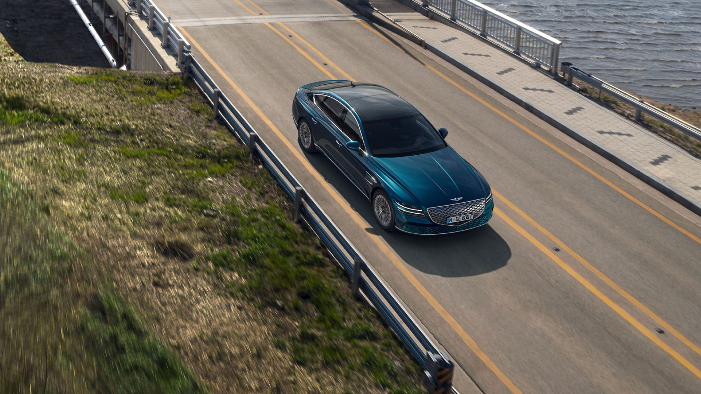 A teal Electrified G80 driving on a bridge, offering a top-down perspective with a river in the background.