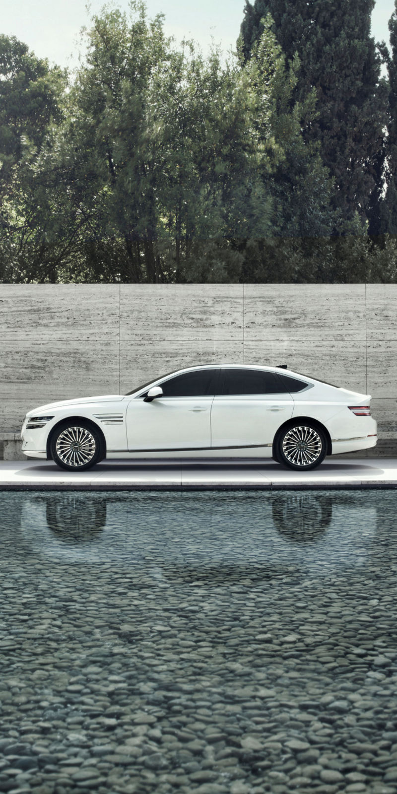 A white Electrified G80 parked near a serene pool with a minimalist concrete wall and lush greenery in the background.