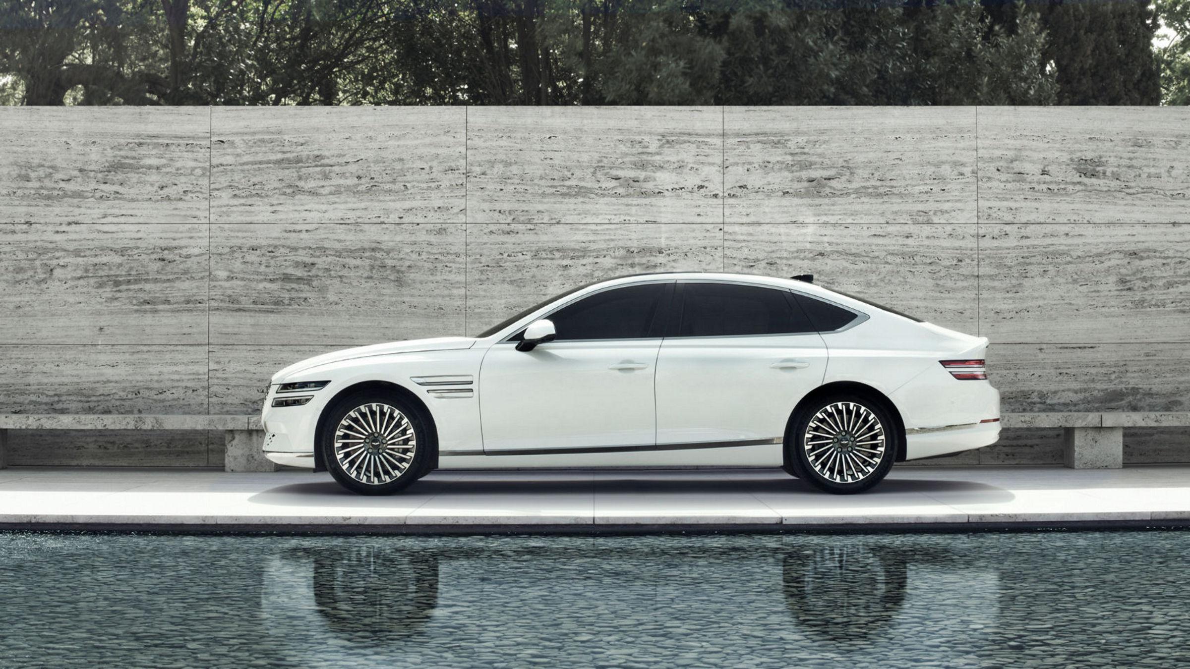 A white Electrified G80 parked near a serene pool with a minimalist concrete wall and lush greenery in the background.