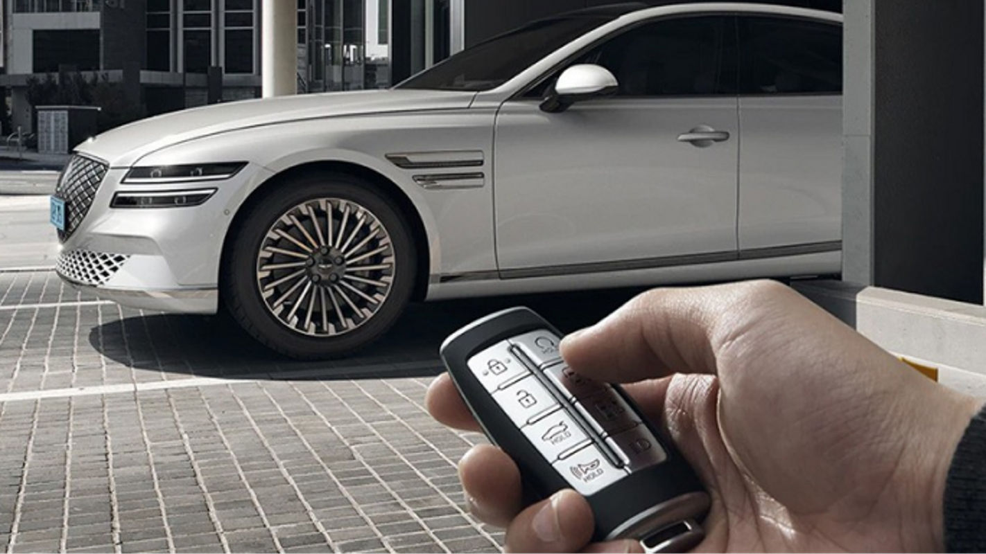 A white Electrified G80 and a gray Electrified G80 parked in a lot, with a key fob being operated in the foreground.