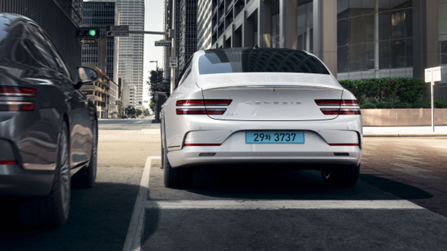  A white Electrified G80 parked at the roadside in an urban area, with a gray sedan nearby.