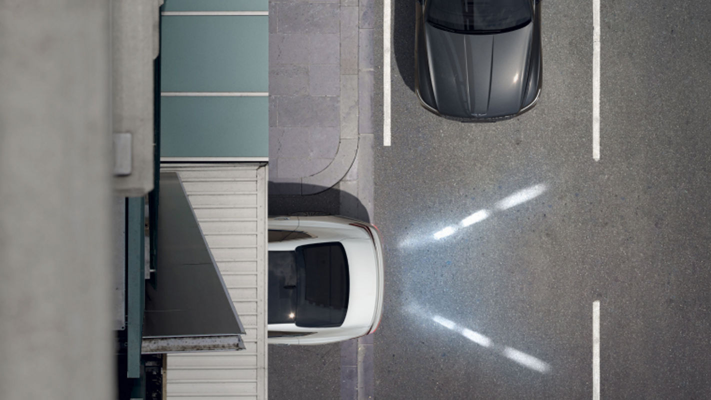 A white Electrified G80 reversing into a narrow parking space with its rear lights and sensors active.