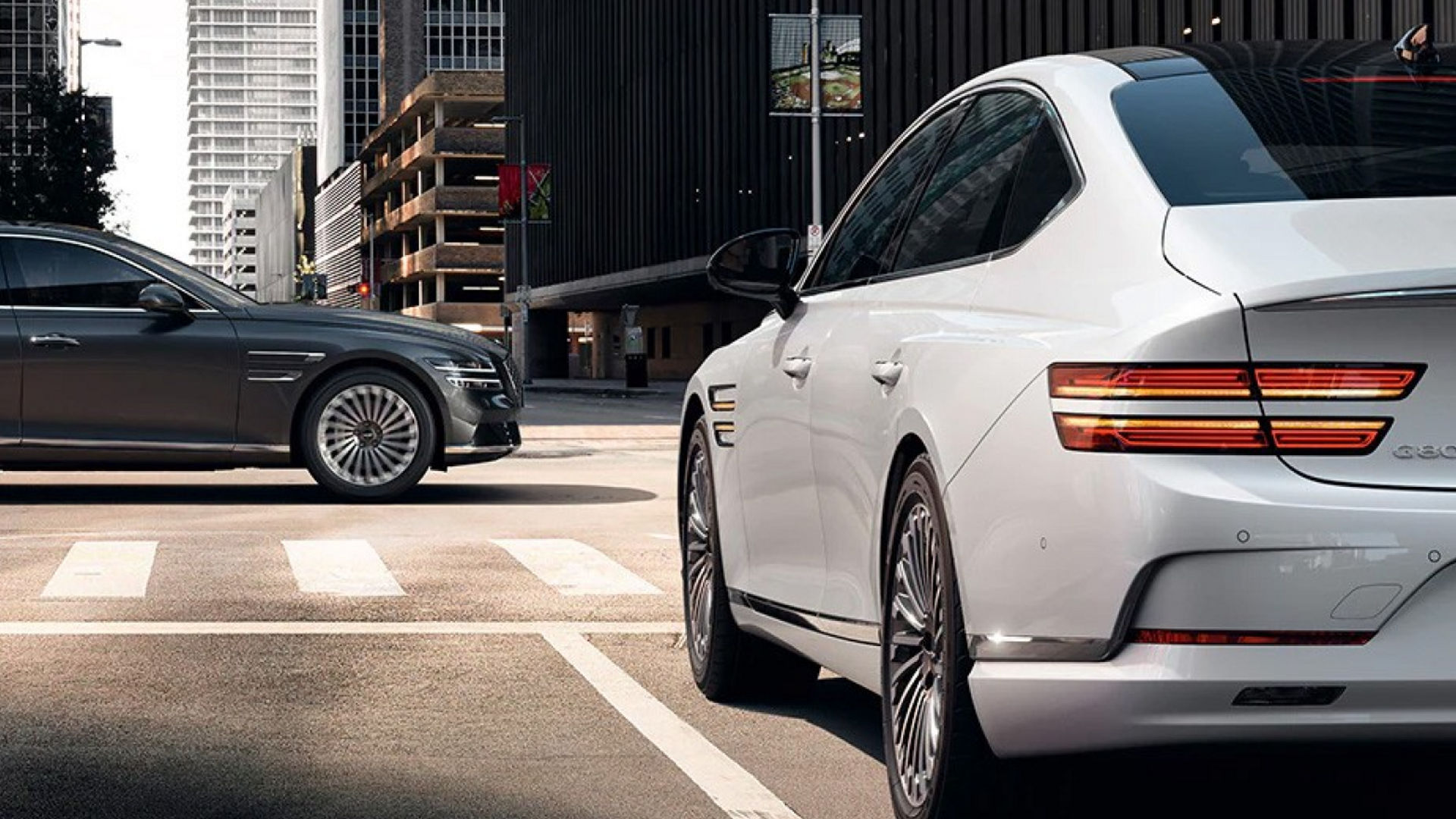 A white Electrified G80 stopped at an urban intersection with a gray sedan in the background.