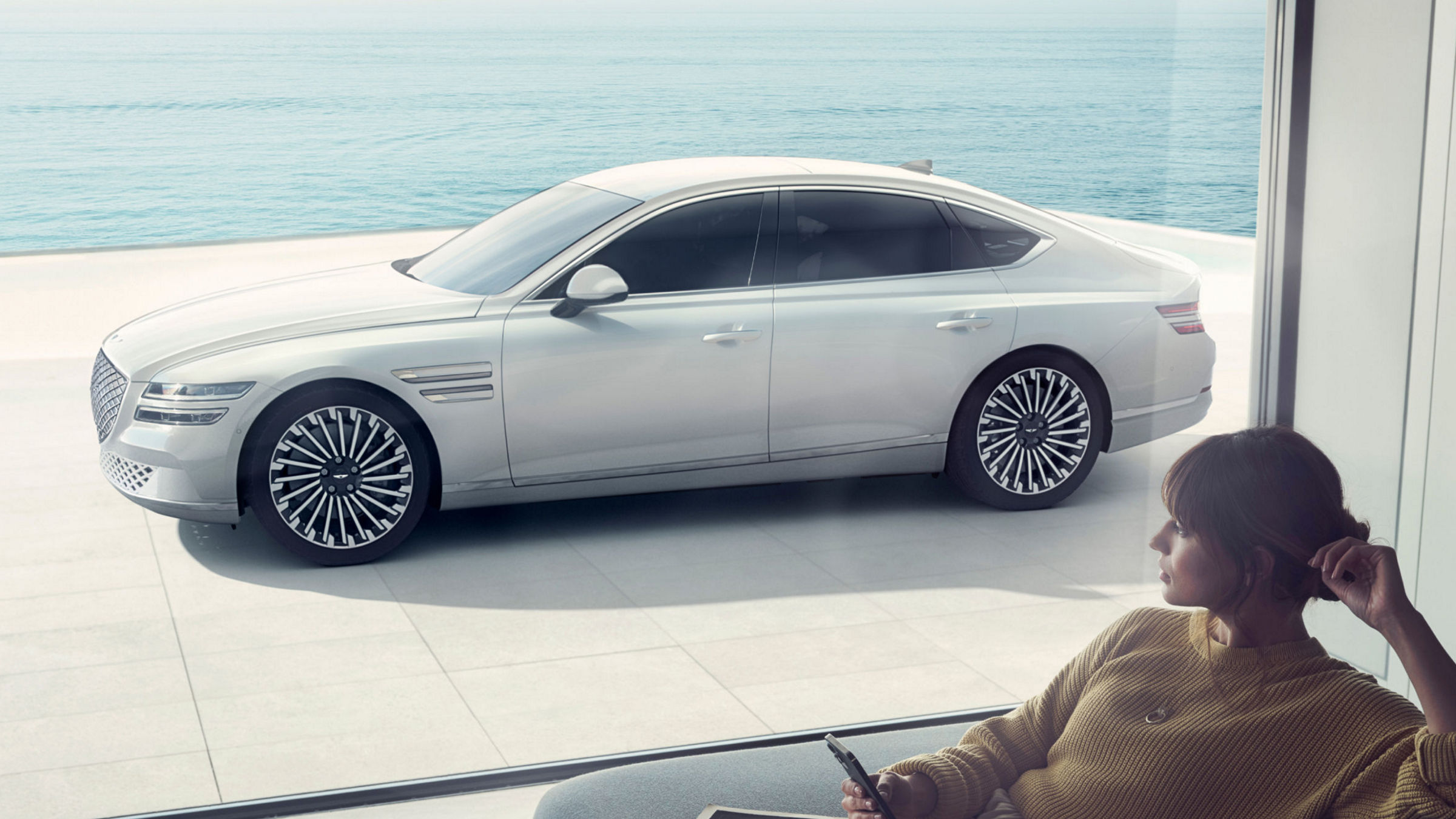 A silver Electrified G80 parked near a coastal setting with a woman sitting indoors, enjoying the view.