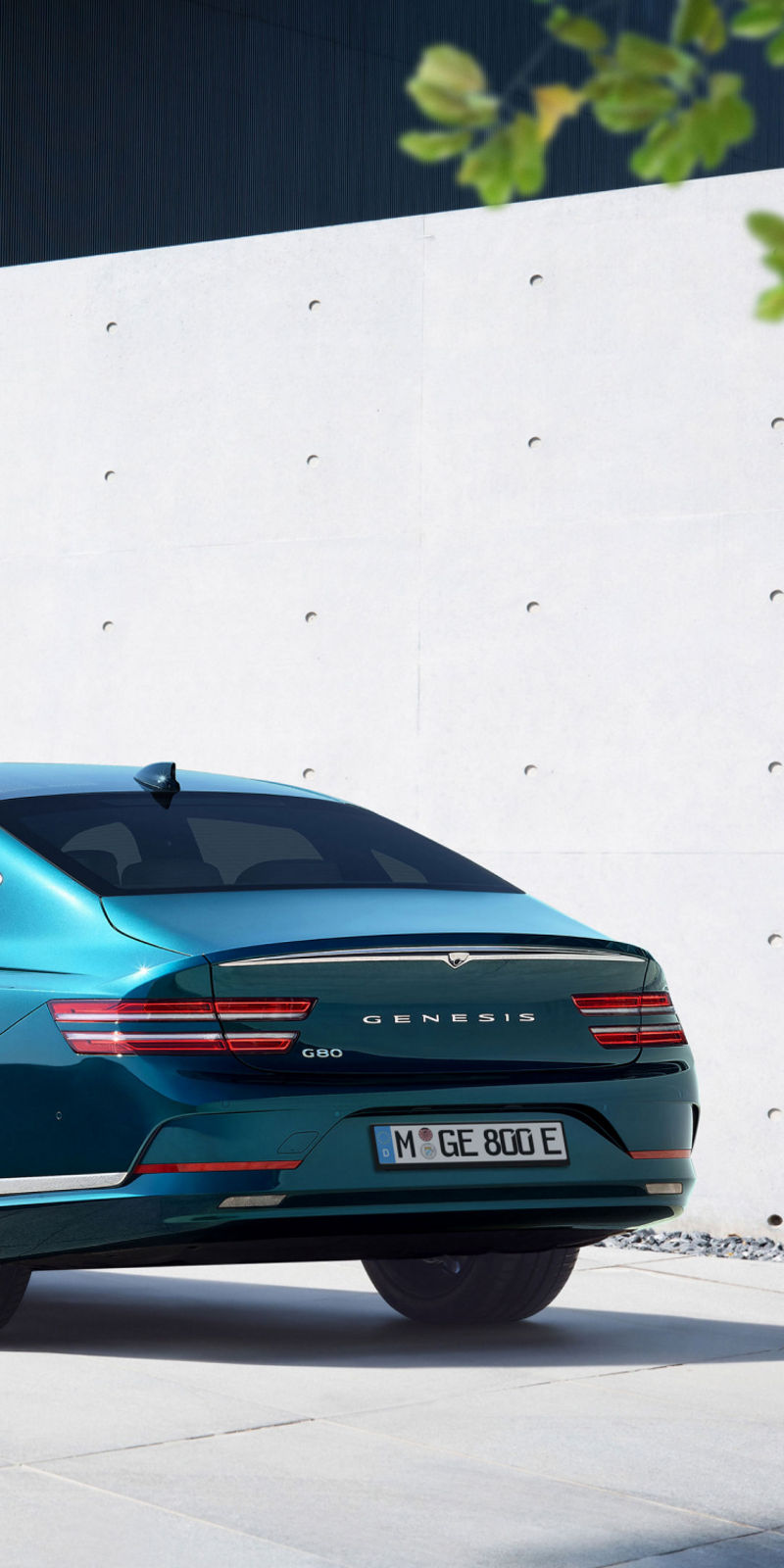 A teal Electrified G80 parked in a modern courtyard with clean architectural elements and soft shadows.