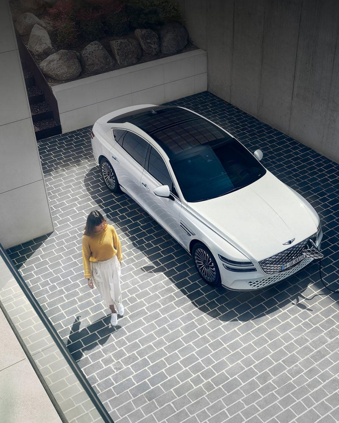 A top-down view of a white Electrified G80 parked on a tiled driveway, with a person walking nearby.