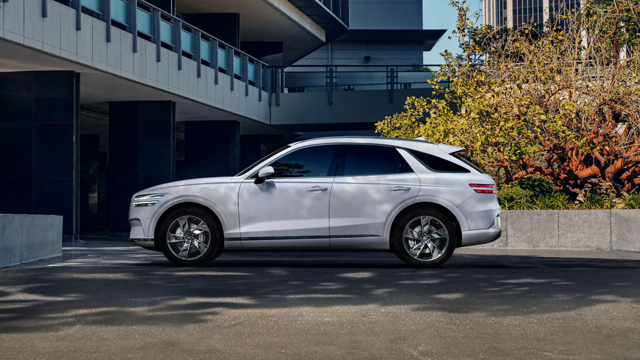 Ein Seitenprofil des Genesis Electrified GV70, der in einer modernen urbanen Umgebung geparkt ist, das sein schlankes Design und die silberne Lackierung hervorhebt