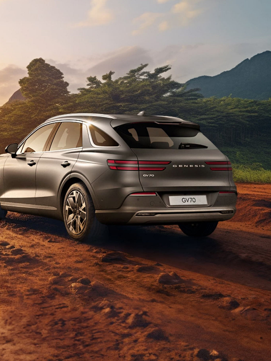 A rear three-quarter view of the Genesis GV70 driving on a rugged dirt road with a scenic mountainous backdrop.