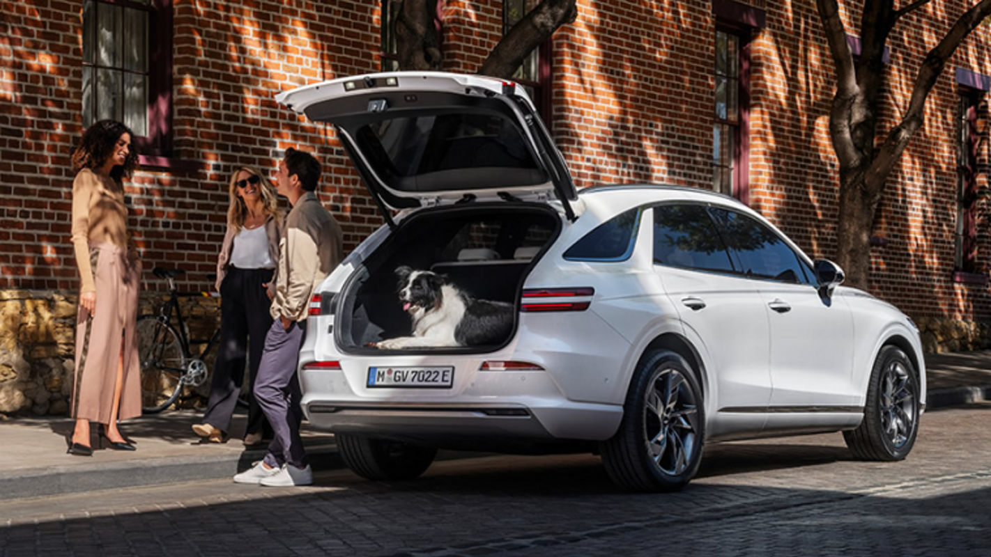 An Electrified Genesis GV70 with its rear hatch open, showing ample cargo space and a pet inside, surrounded by a group of people.