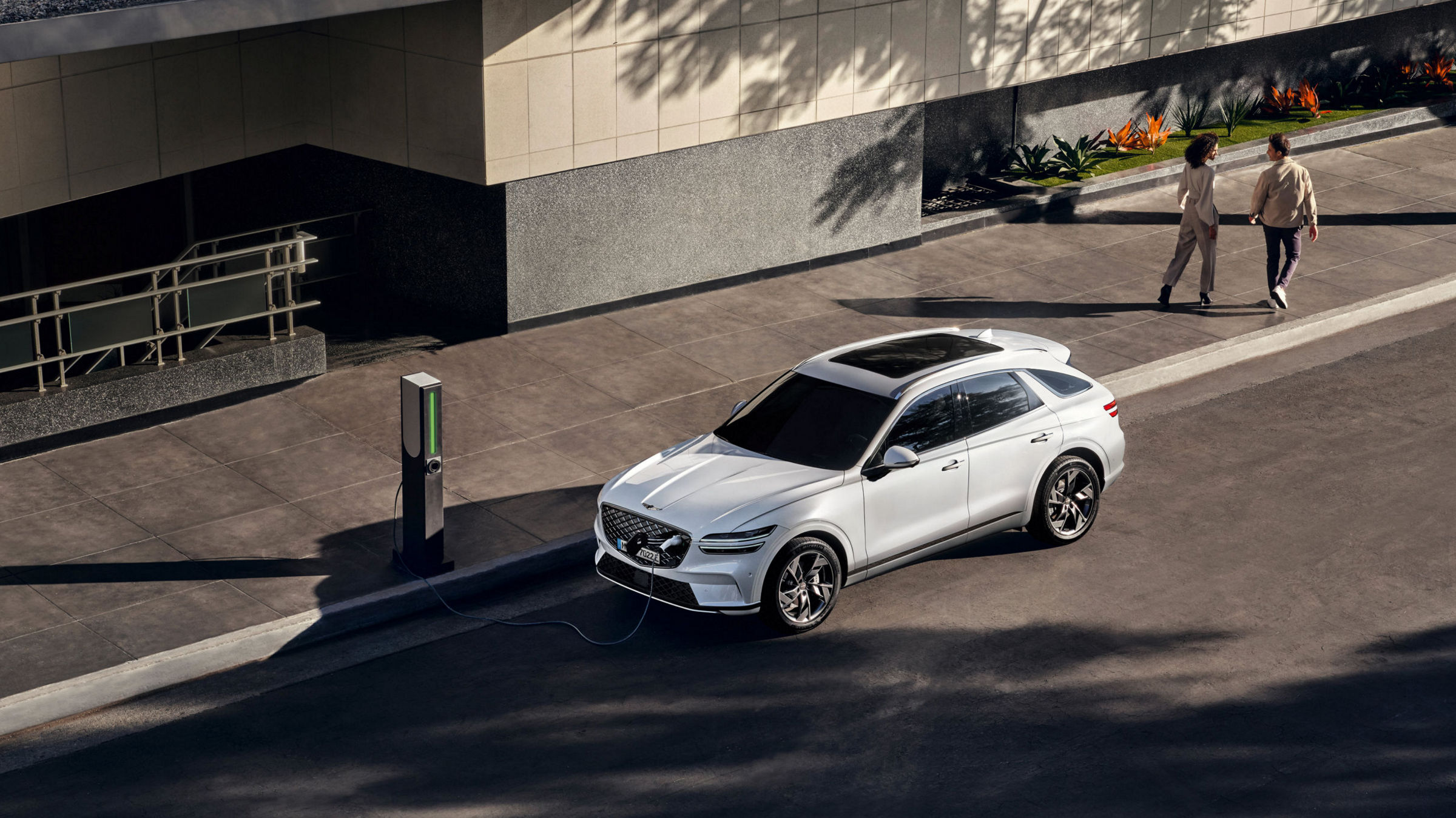 An aerial view of the Genesis Electrified GV70 parked at a public charging station, with two people walking nearby.