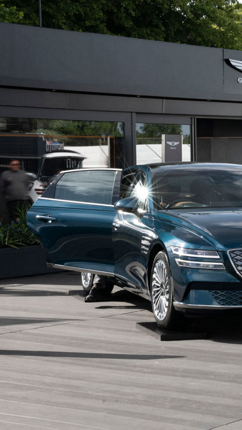 A Genesis Electrified G80 sedan is displayed at an outdoor event, with a showroom booth in the background and blurred people walking by.