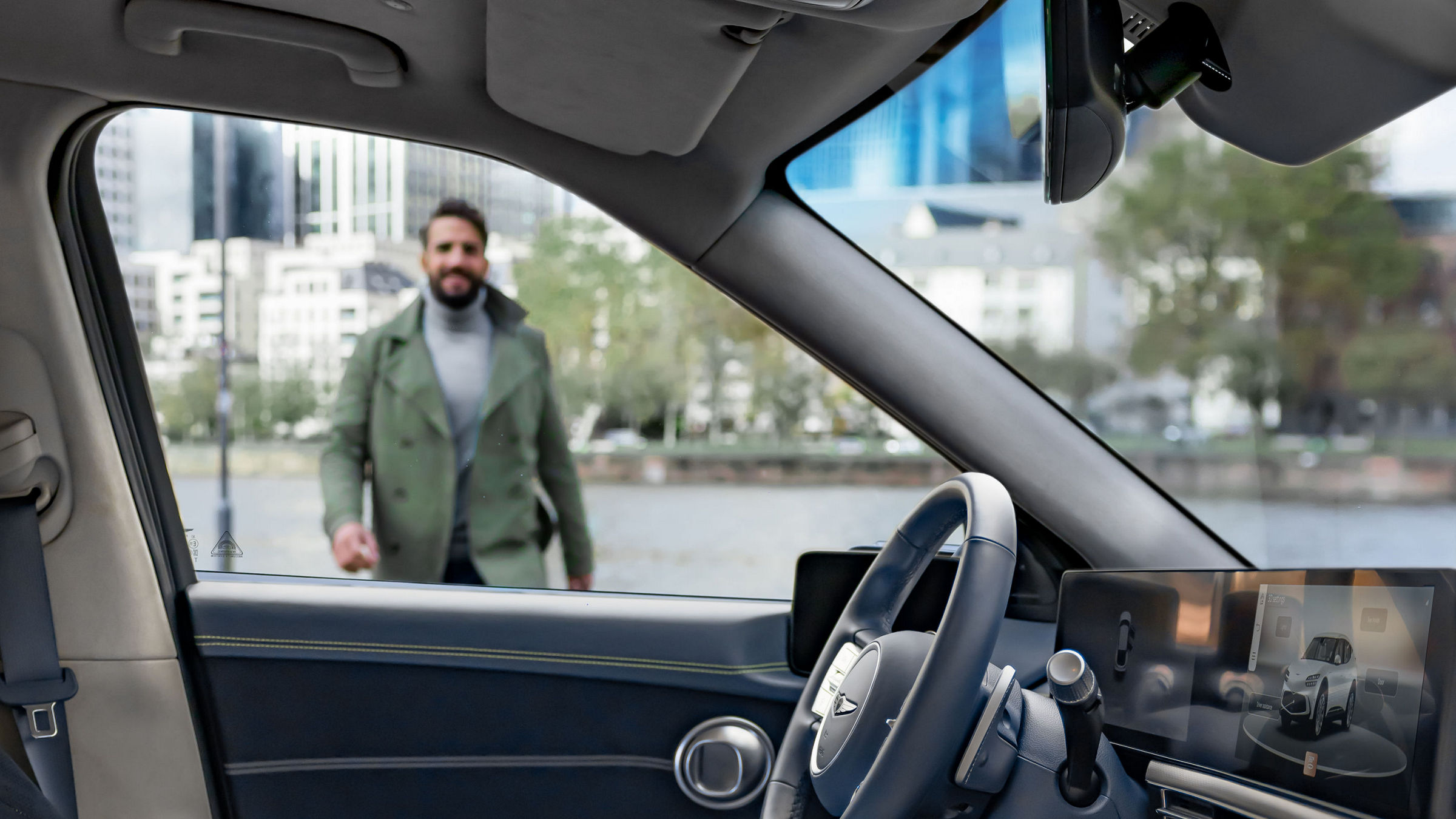 A man in a green jacket is seen outside a modern car, viewed from the driver's seat perspective. The background features a cityscape with tall buildings and trees.