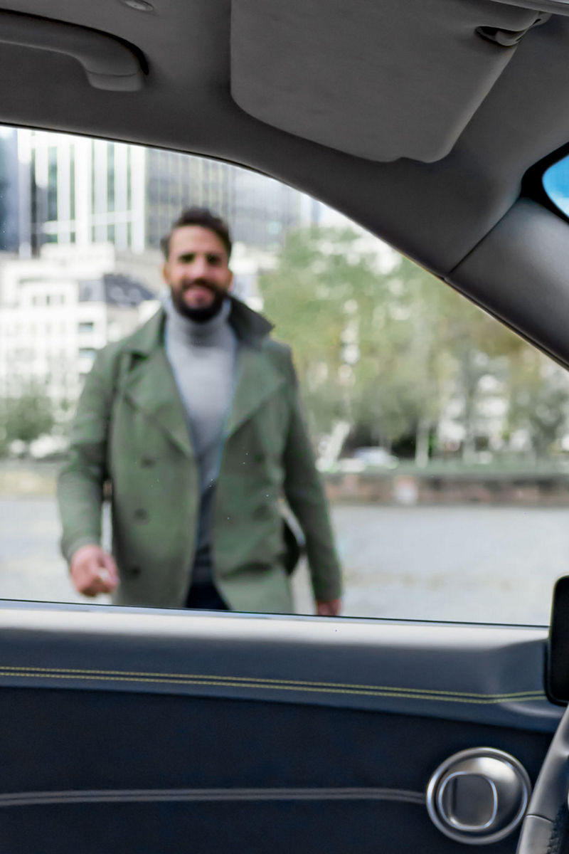 A man in a green jacket is seen outside a modern car, viewed from the driver's seat perspective. The background features a cityscape with tall buildings and trees.