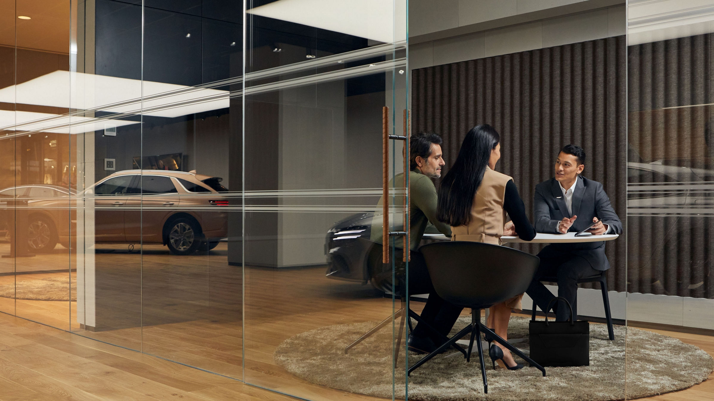 A professional meeting inside a modern glass-walled office, with a Genesis vehicle visible in the background showroom.