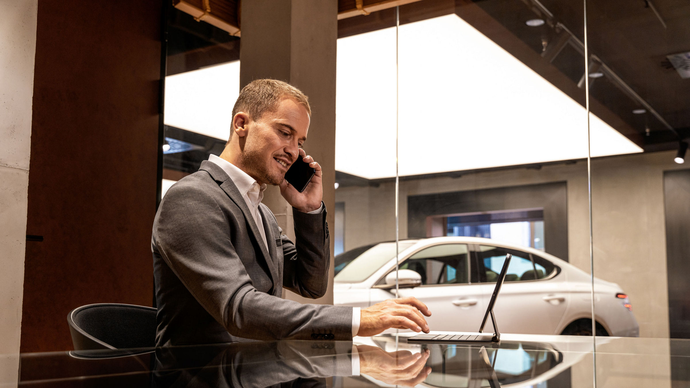 A man dressed in a suit speaks on the phone while working on his laptop, with a Genesis car visible behind the glass wall.