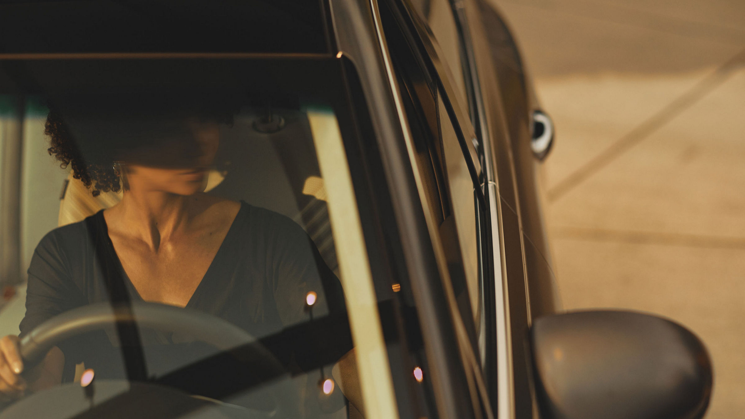 A woman sits confidently behind the wheel of a black Genesis G80, the lighting casting a warm ambiance through the car's sleek windshield.