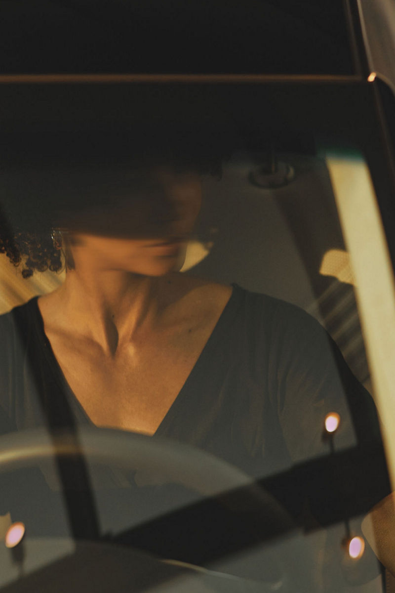 A woman sits confidently behind the wheel of a black Genesis G80, the lighting casting a warm ambiance through the car's sleek windshield.