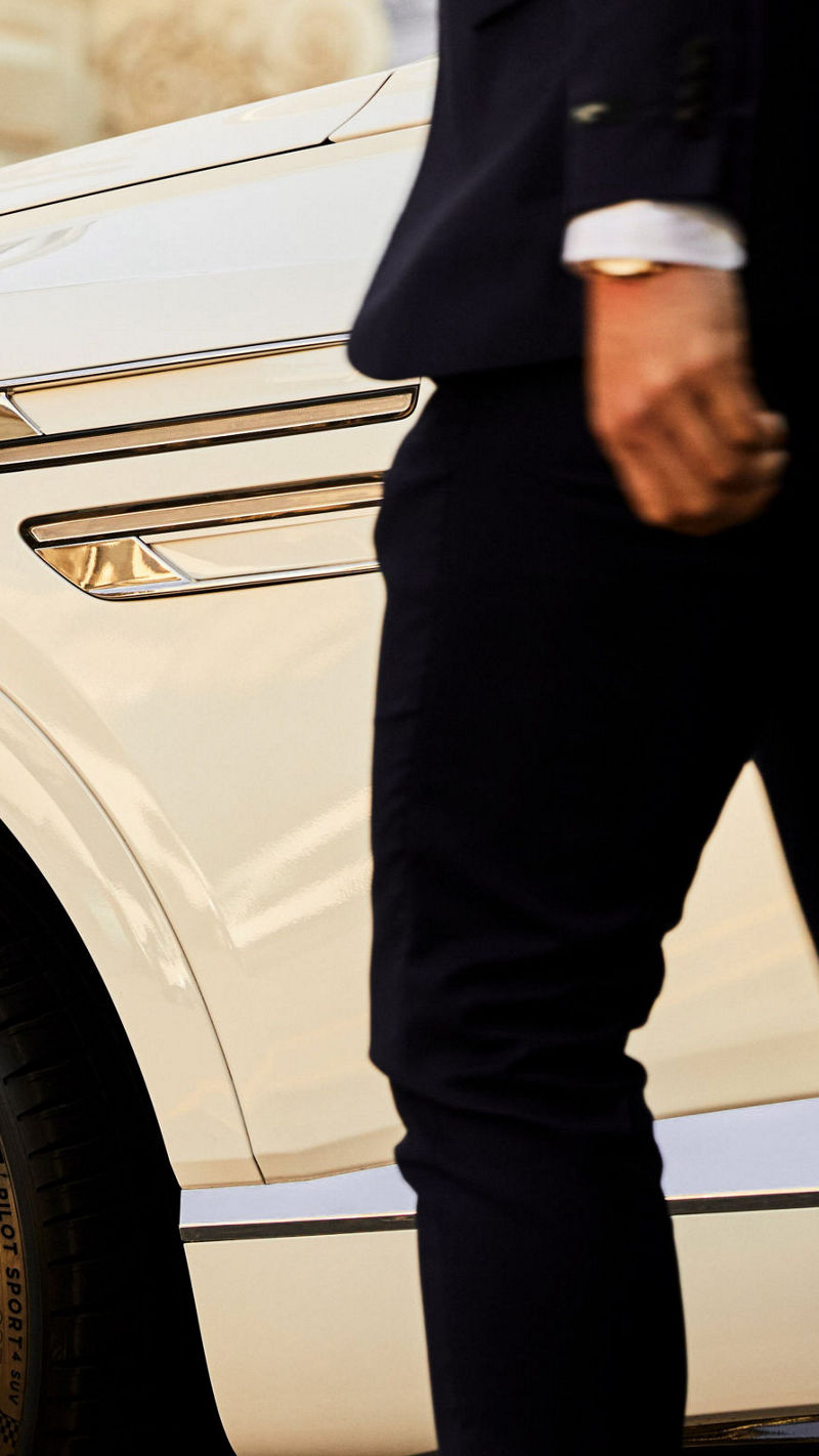 A person in a suit walks past a white Genesis GV80, focusing on the luxurious chrome details and the vehicle's premium alloy wheels.