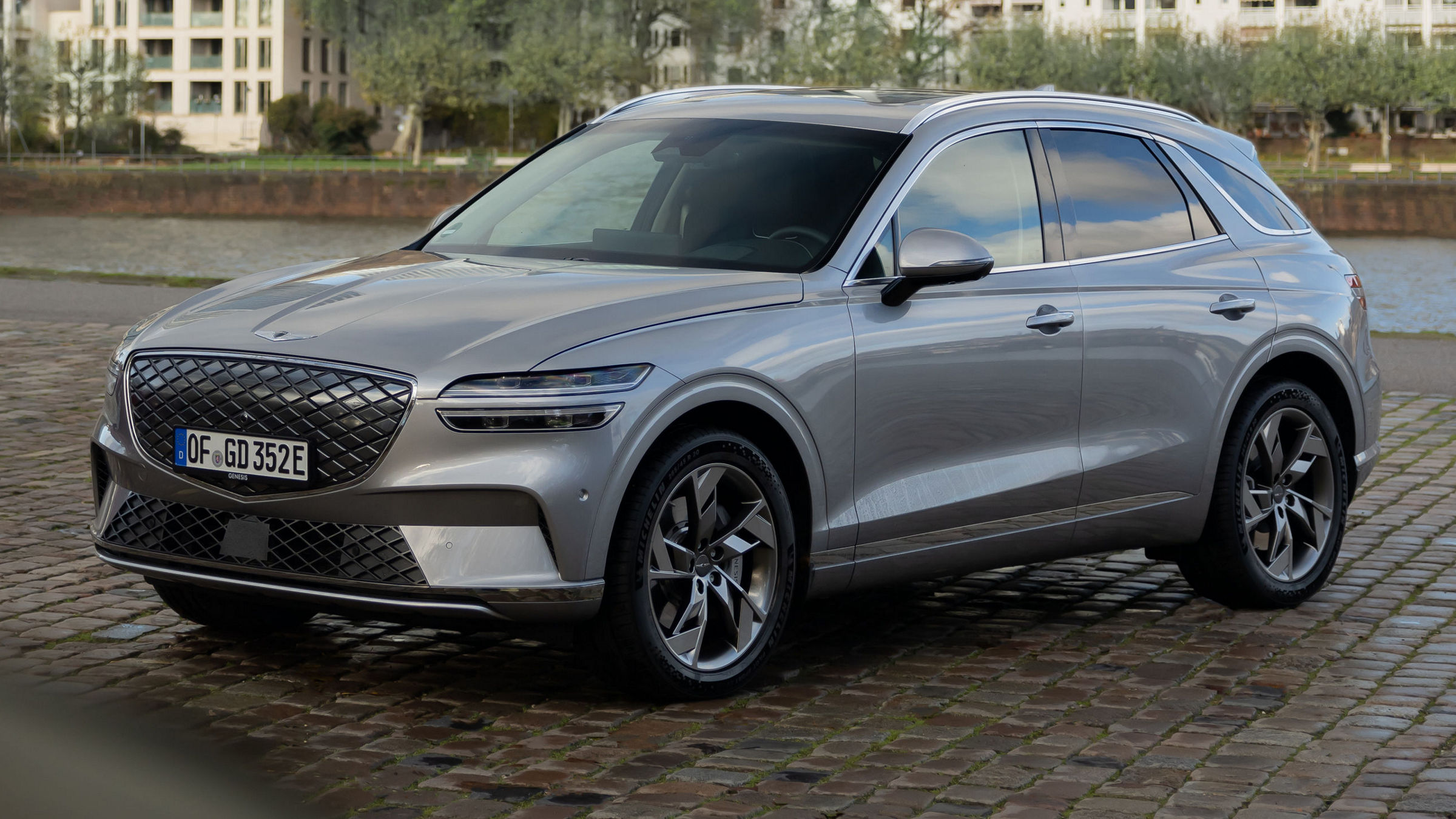 A silver Genesis GV80 is parked near a riverbank with urban apartments in the background, highlighting its elegant exterior design.