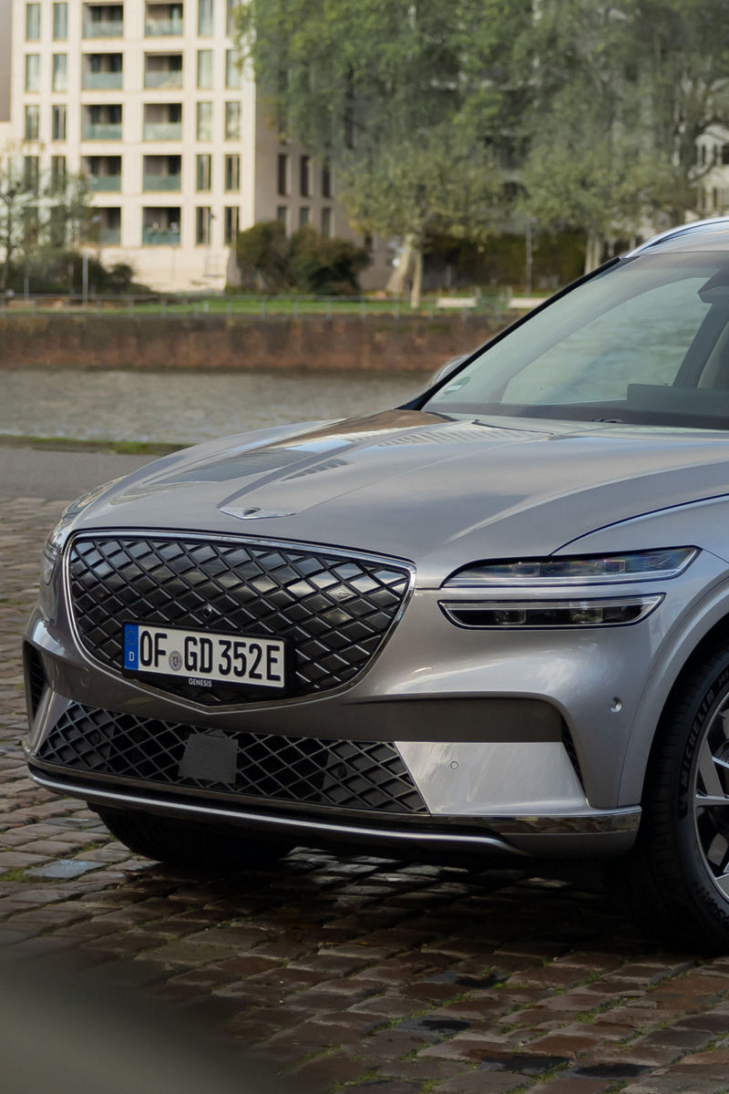 A silver Genesis GV80 is parked near a riverbank with urban apartments in the background, highlighting its elegant exterior design.