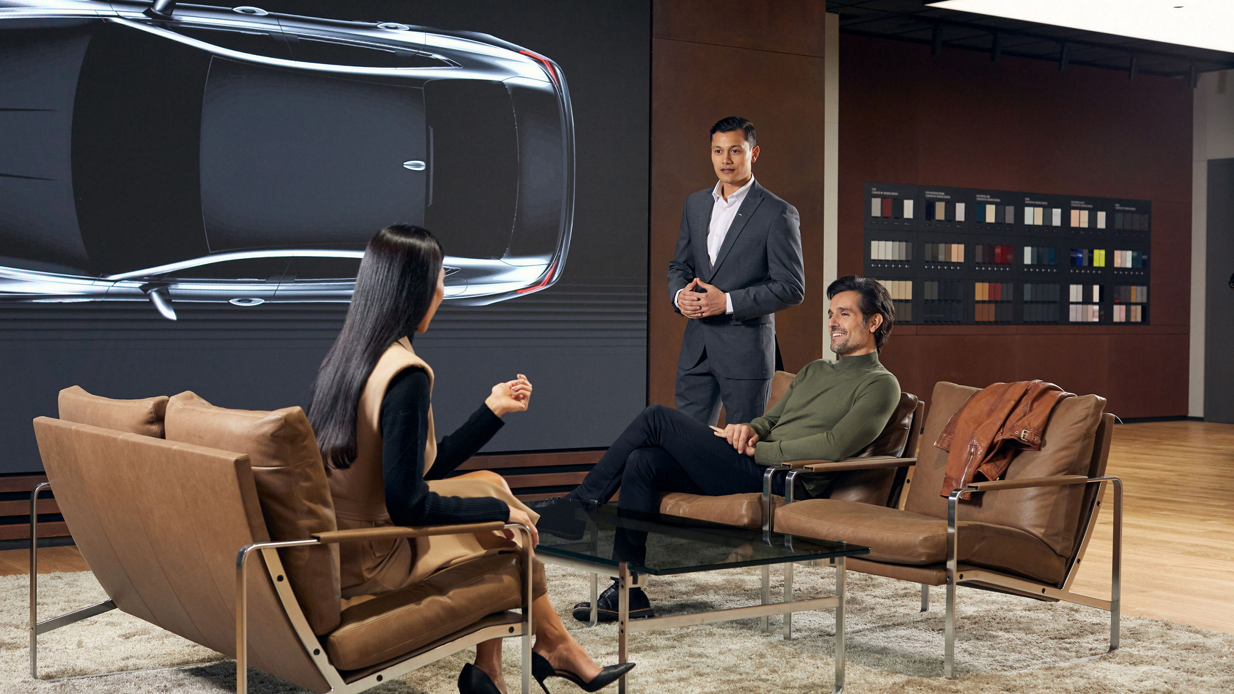  A professional discussion is taking place, with a man presenting to two seated individuals, a car design and material options displayed on the wall in the background.