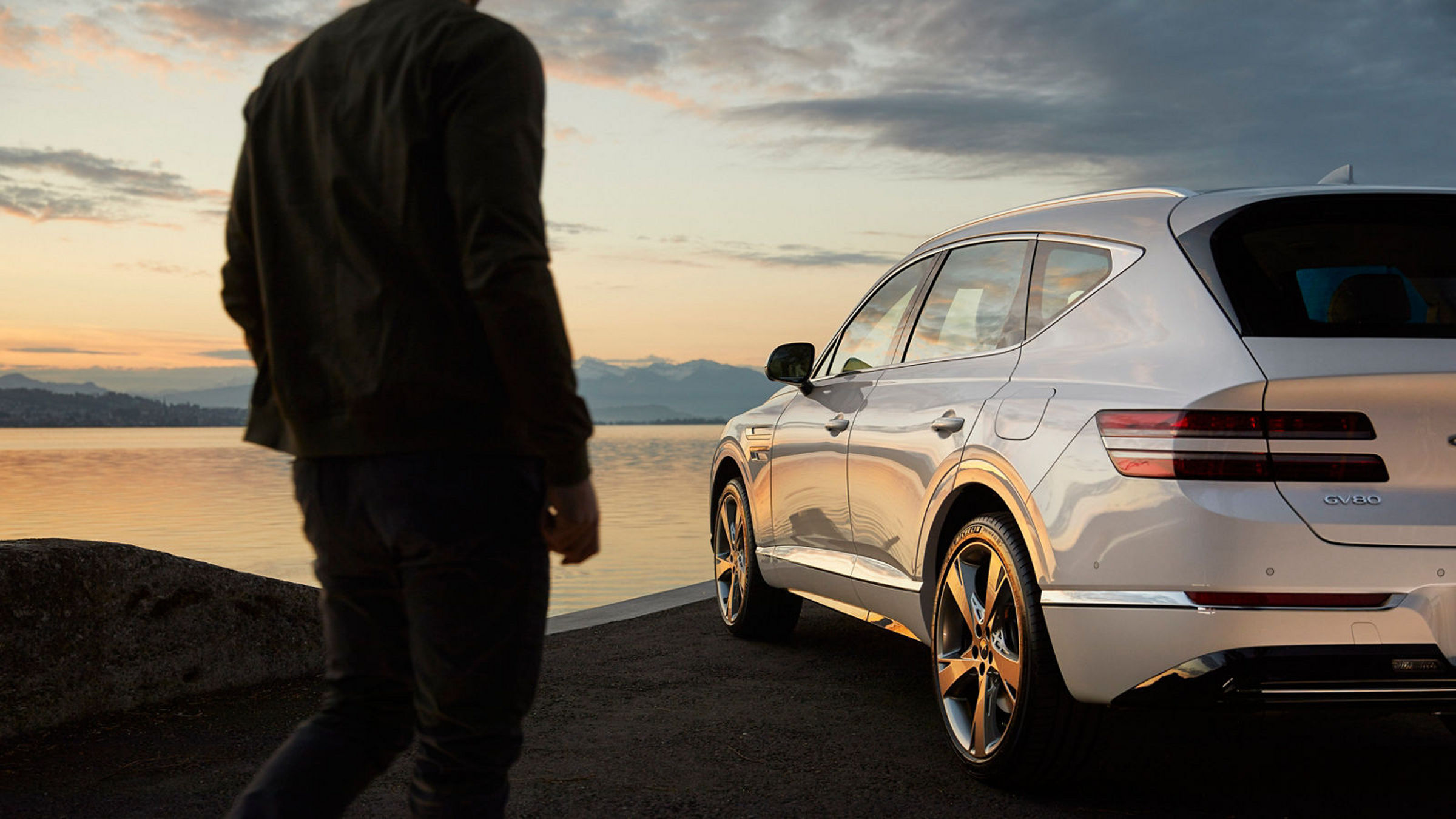 A man walks toward a Genesis GV80 parked beside a scenic lake during sunset, emphasizing the car's luxurious rear design.