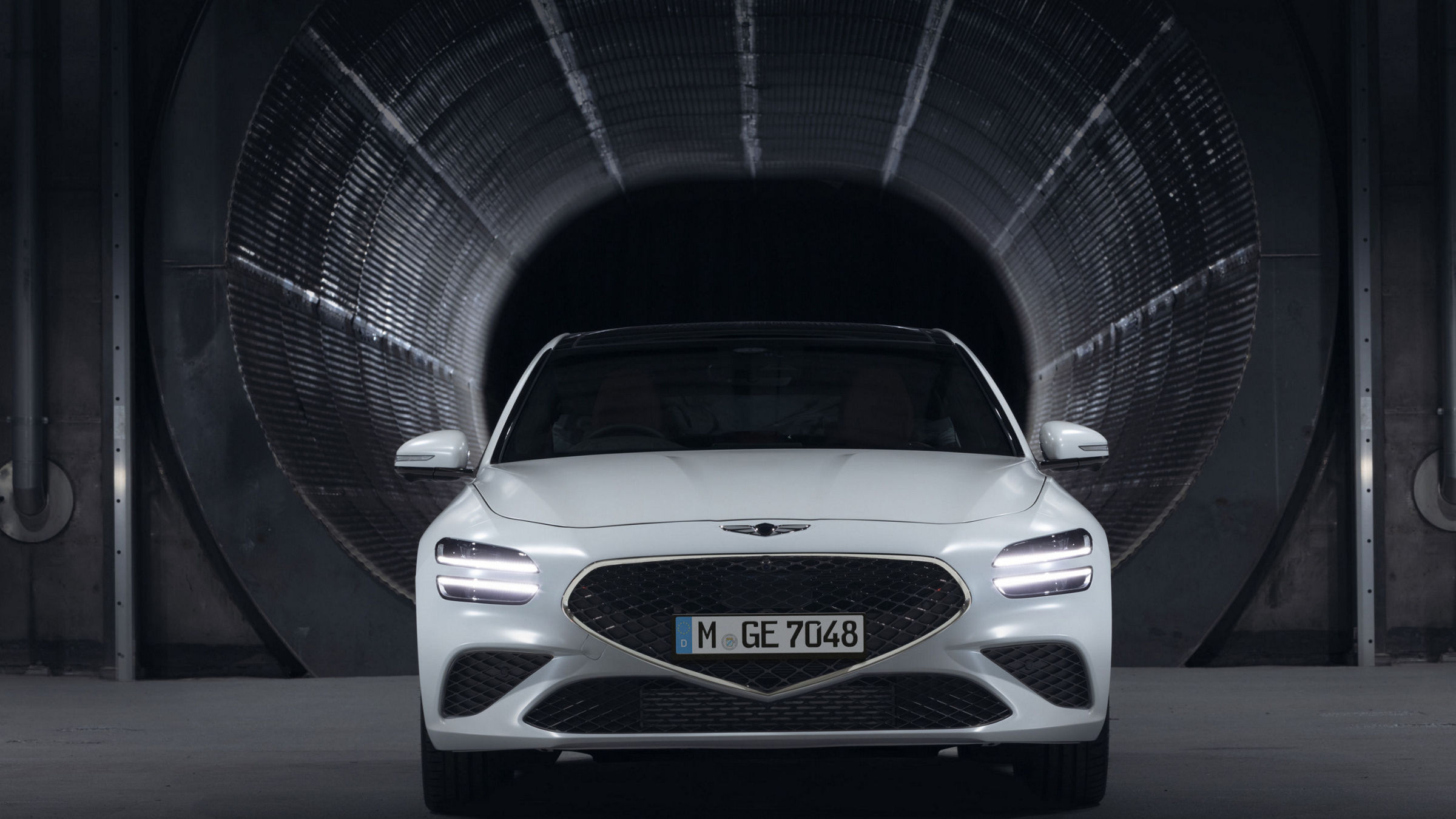 A white luxury car is positioned in front of a dark tunnel, with its headlights on. The sleek design and emblem suggest a high-end model.