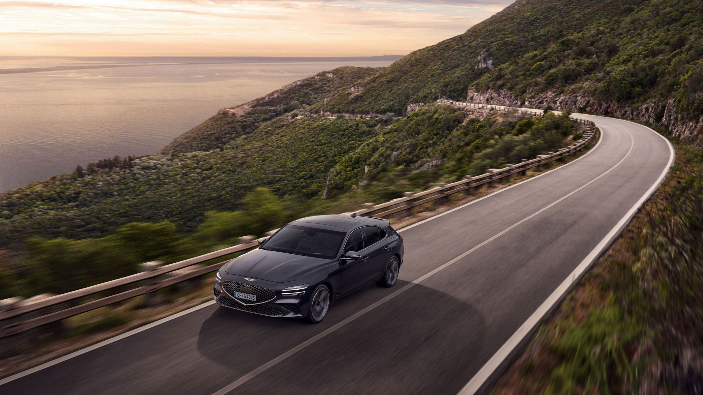 A Genesis G70 drives on a road in the mountains with the sea in the background.