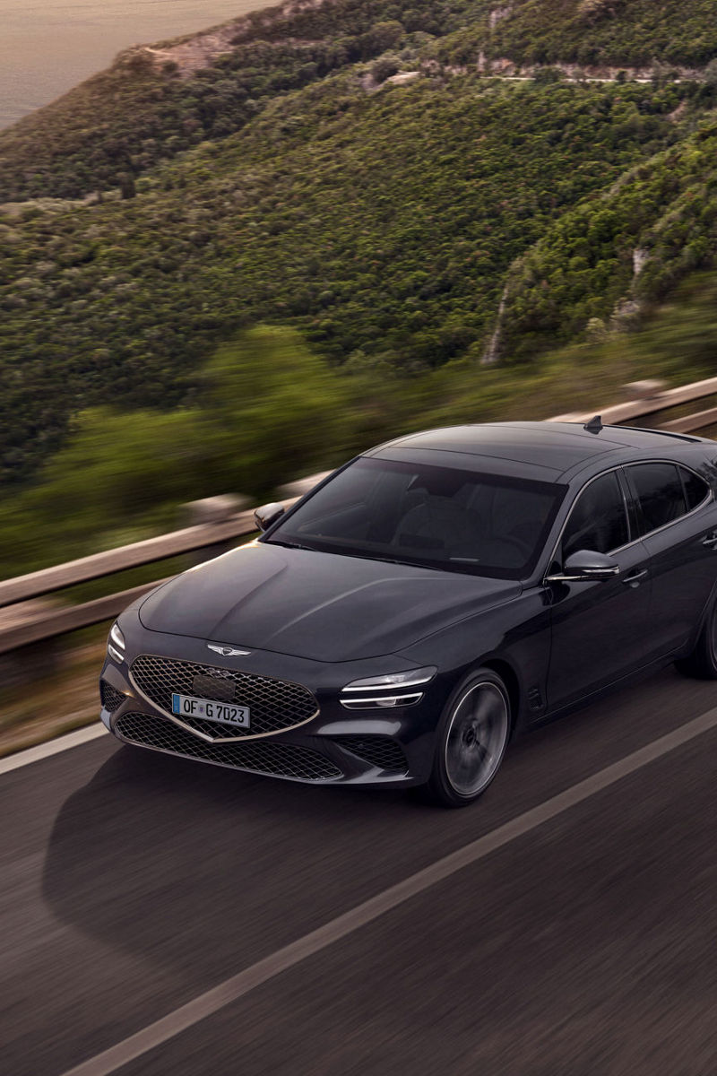 A Genesis G70 drives on a road in the mountains with the sea in the background.