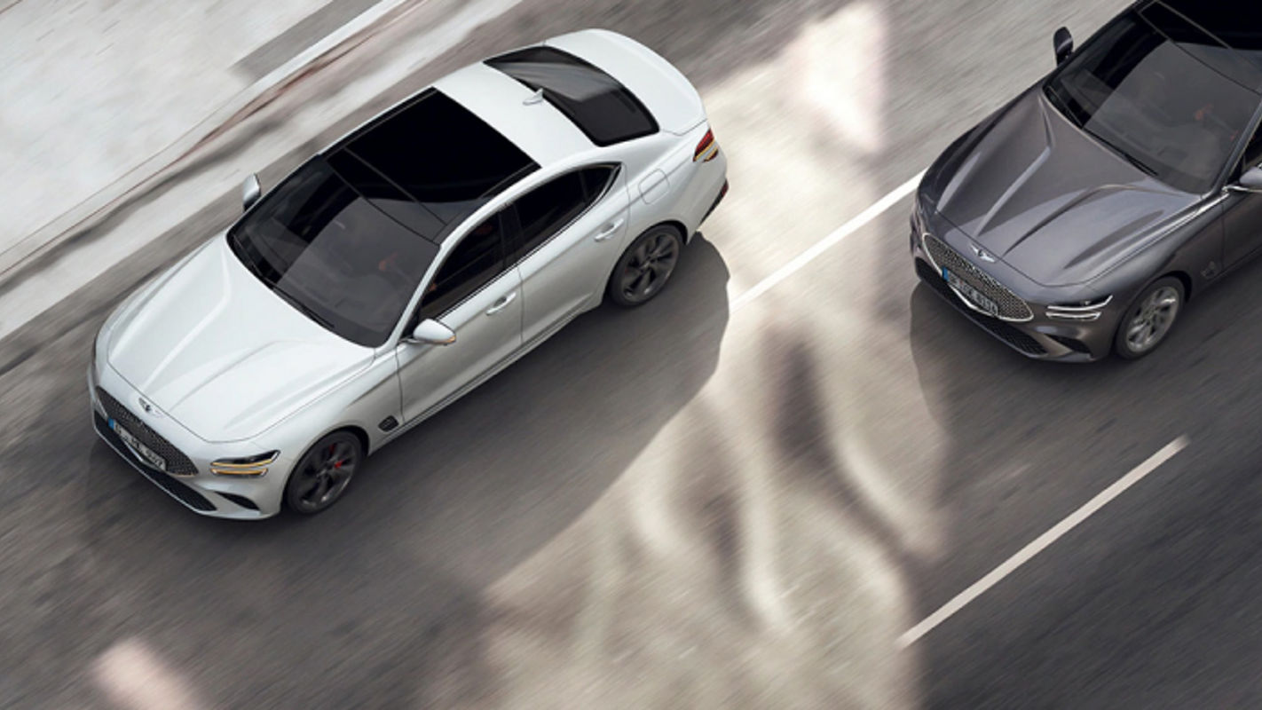 A white Genesis G70 overtaking a gray Genesis G70 on a city street, viewed from above.