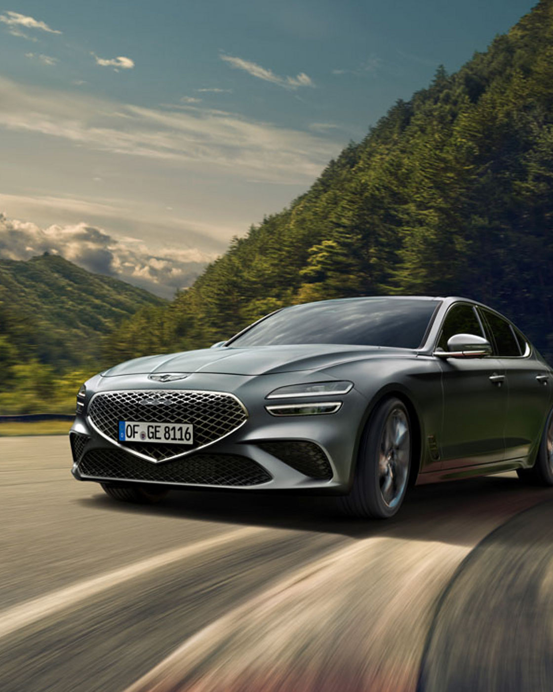 A Genesis G70 driving on a winding road with forested hills in the background.