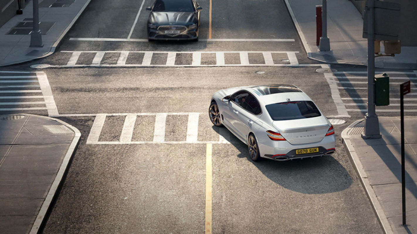 A white Genesis G70 driving through a city intersection in daylight.