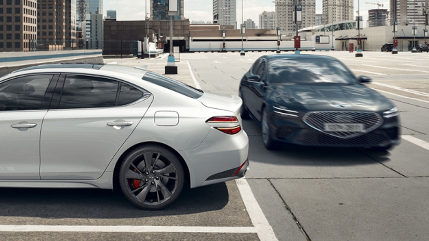 A white Genesis G70 parked in a rooftop parking lot with a dark gray G70 passing nearby.