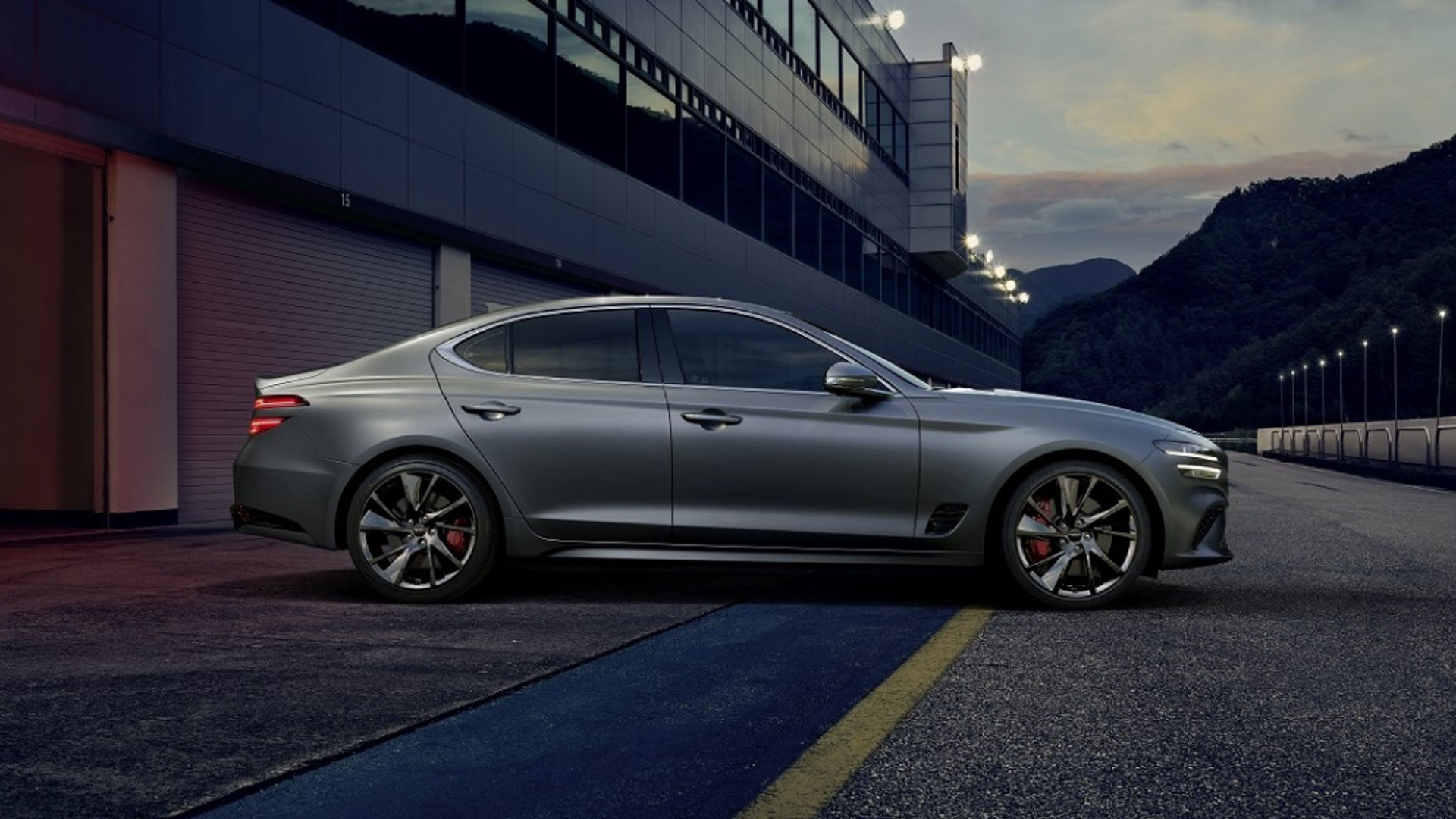 A silver Genesis G70 parked on a race track at dusk with mountains in the background.