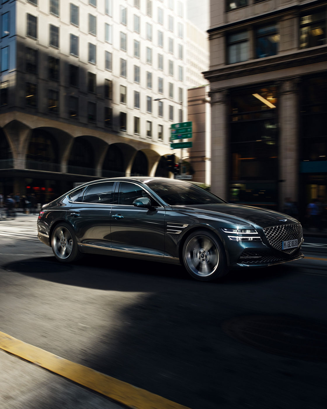 Genesis G80 in blue, captured outdoors on a road with people in the background, showing a front-side view.