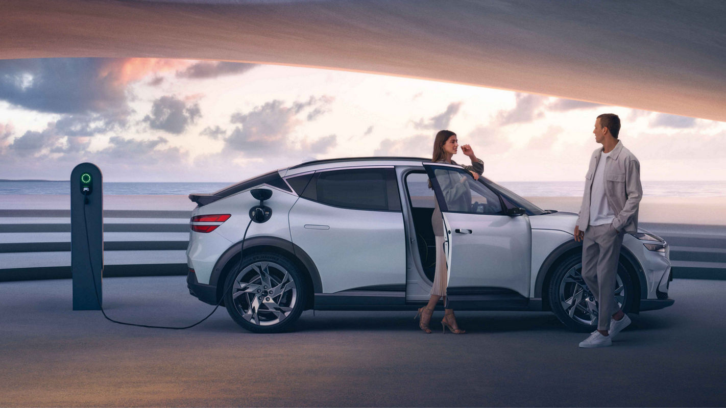 A modern electric car being charged at a charging station, with a stylishly dressed man and woman standing near the vehicle under a futuristic canopy at sunset.