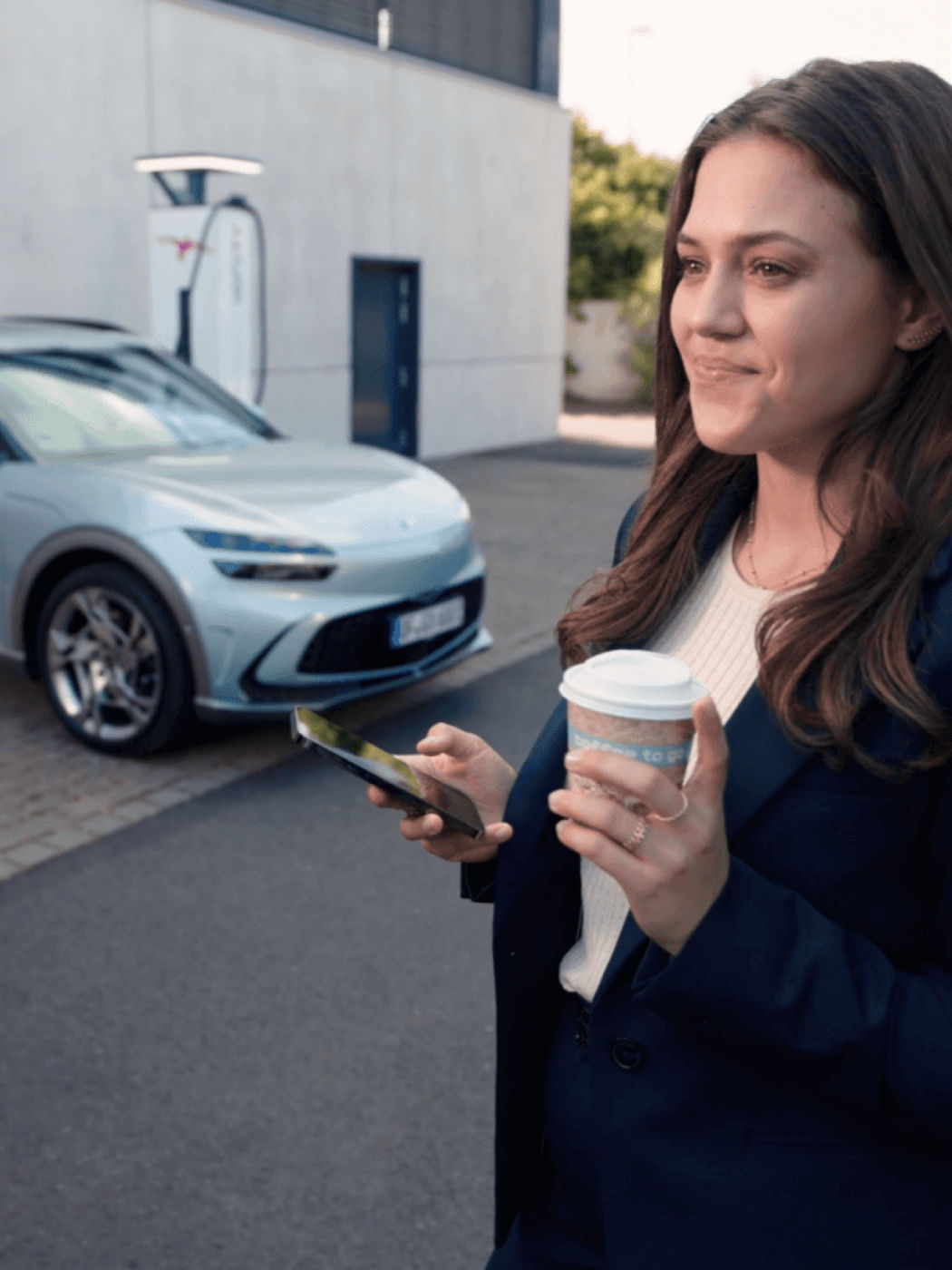 A person holding a coffee cup and smartphone near a GV60 at a charging station