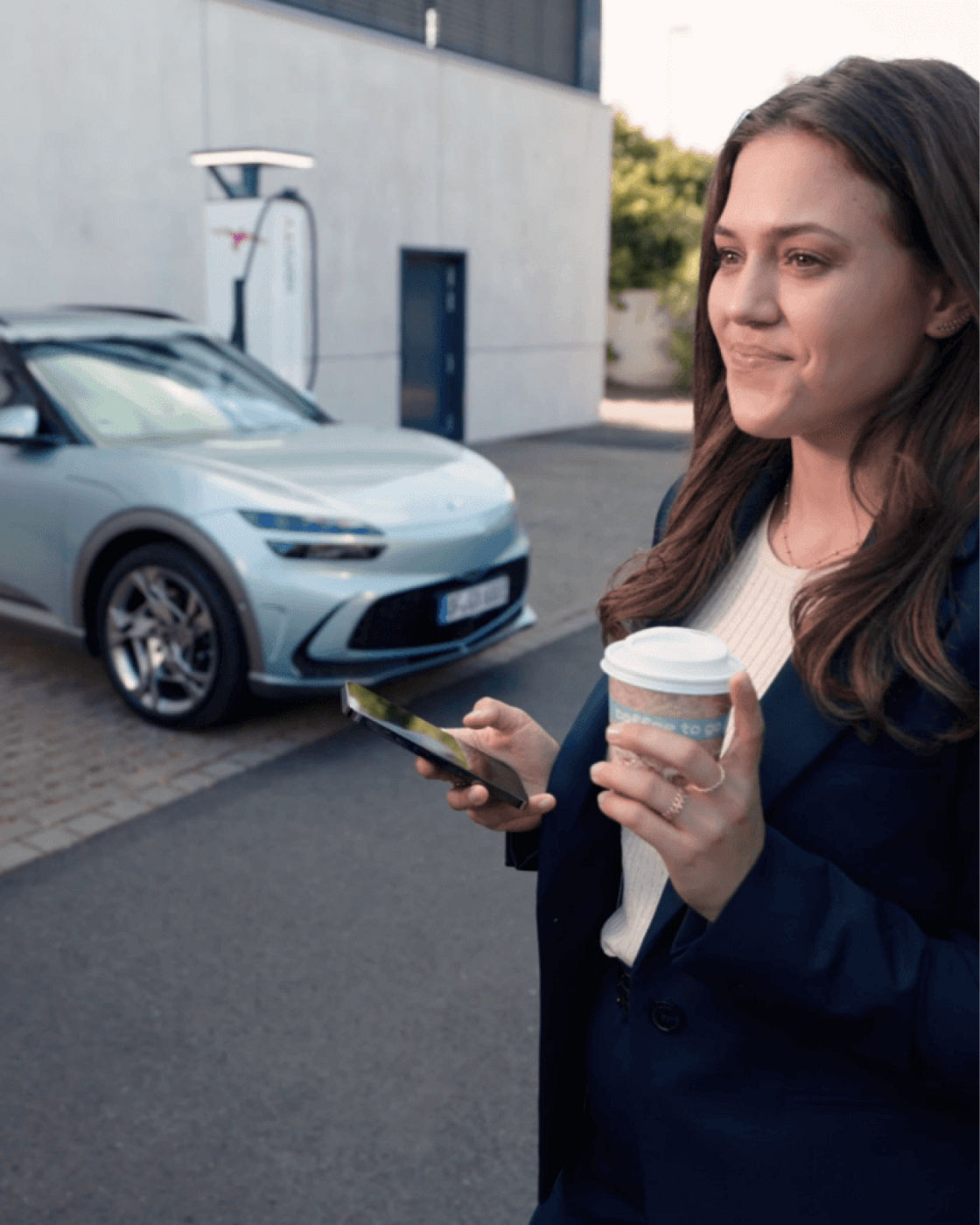 A person holding a coffee cup and smartphone near a GV60 at a charging station