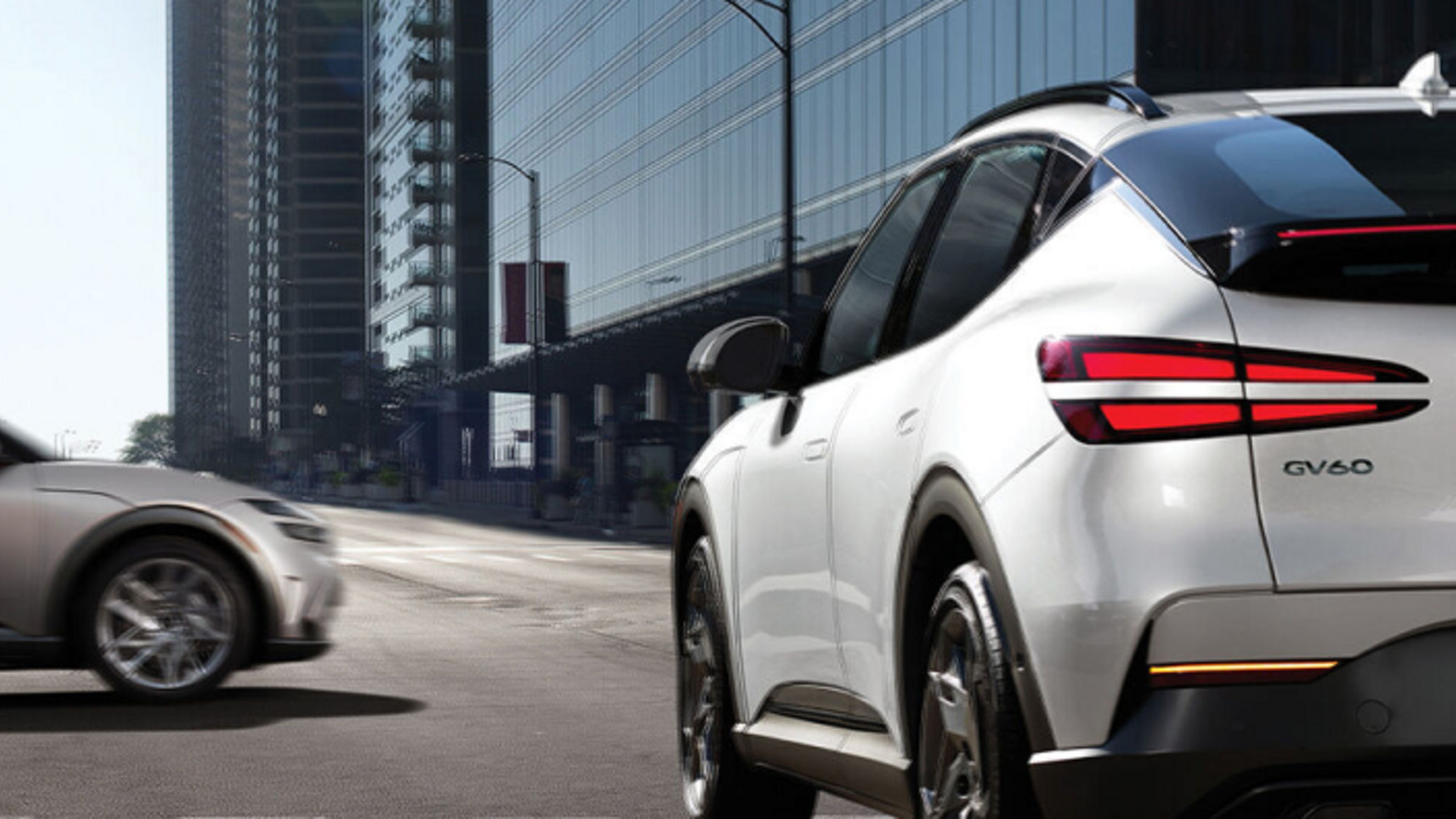 The GV60 waiting at a crosswalk, stopping at the traffic signal, with a silver car passing by next to it.