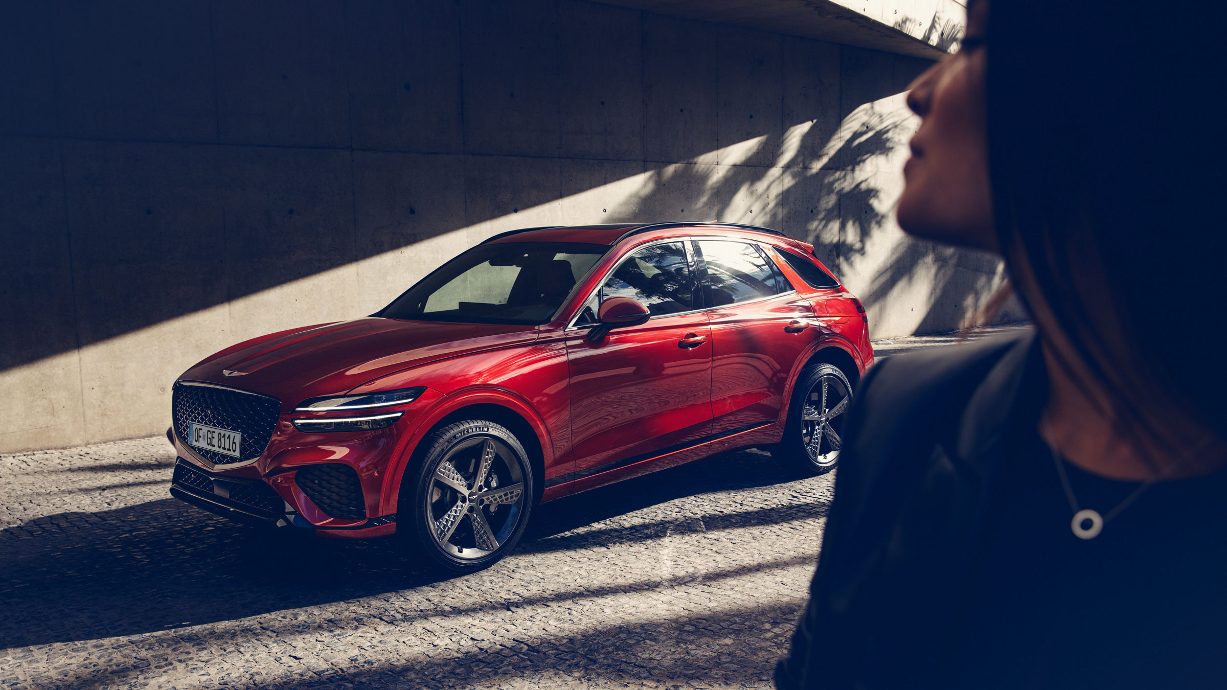 A striking red Genesis GV70 parked under modern architecture, with a woman in the foreground.