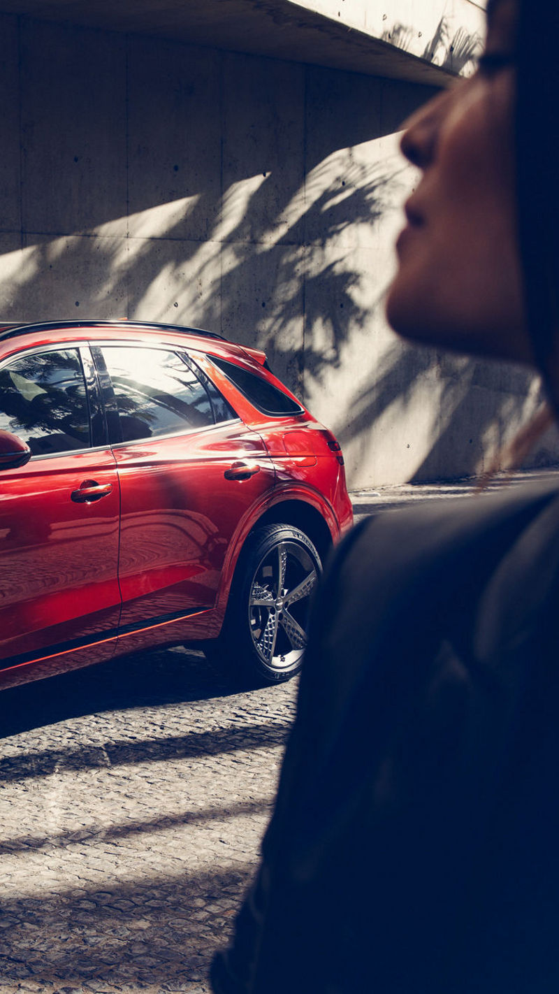 A striking red Genesis GV70 parked under modern architecture, with a woman in the foreground.