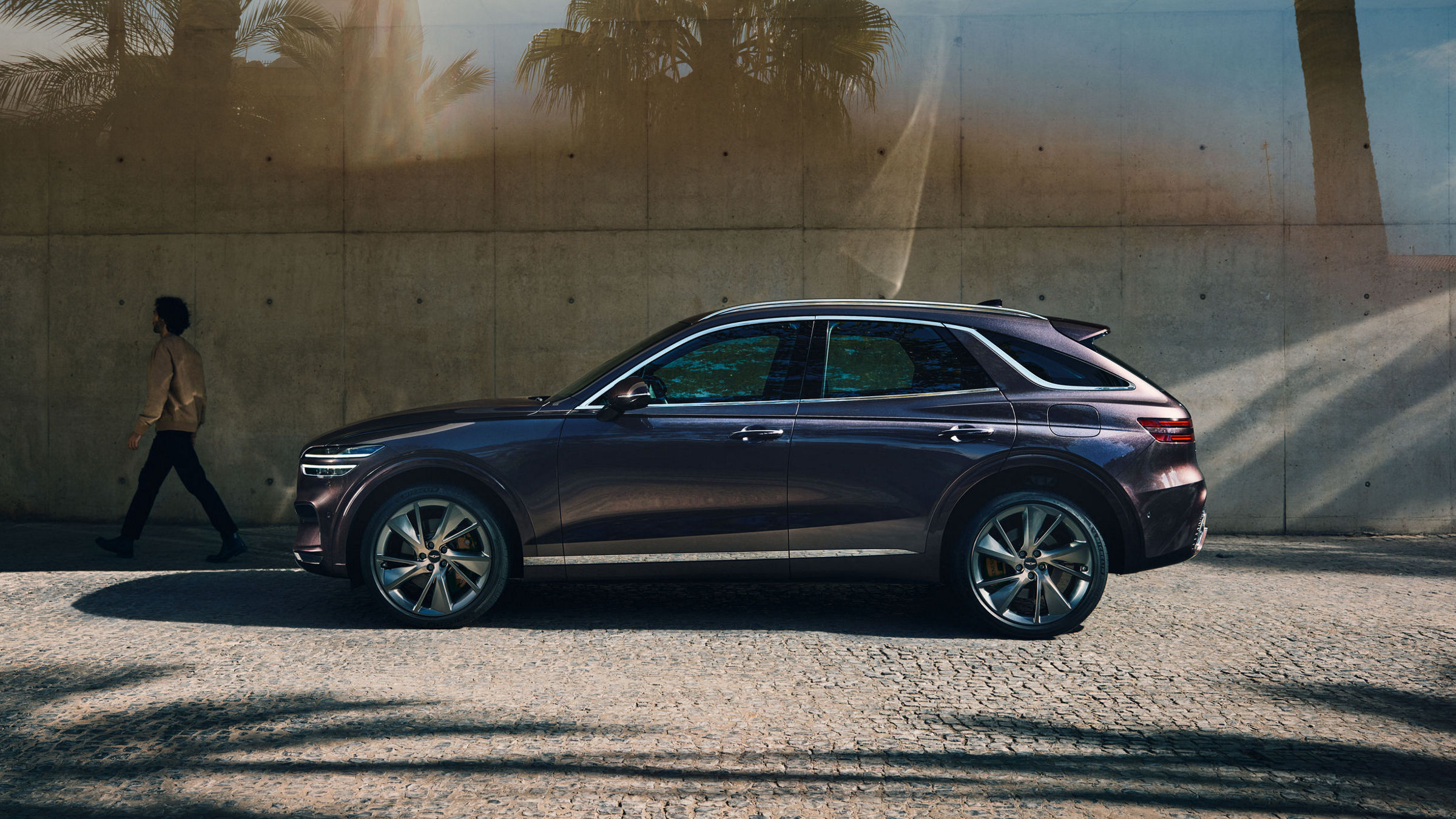 The Genesis GV70 is parked on a cobblestone street beside a modern concrete wall, with palm trees reflecting in the glass, emphasizing its sleek and sophisticated profile.