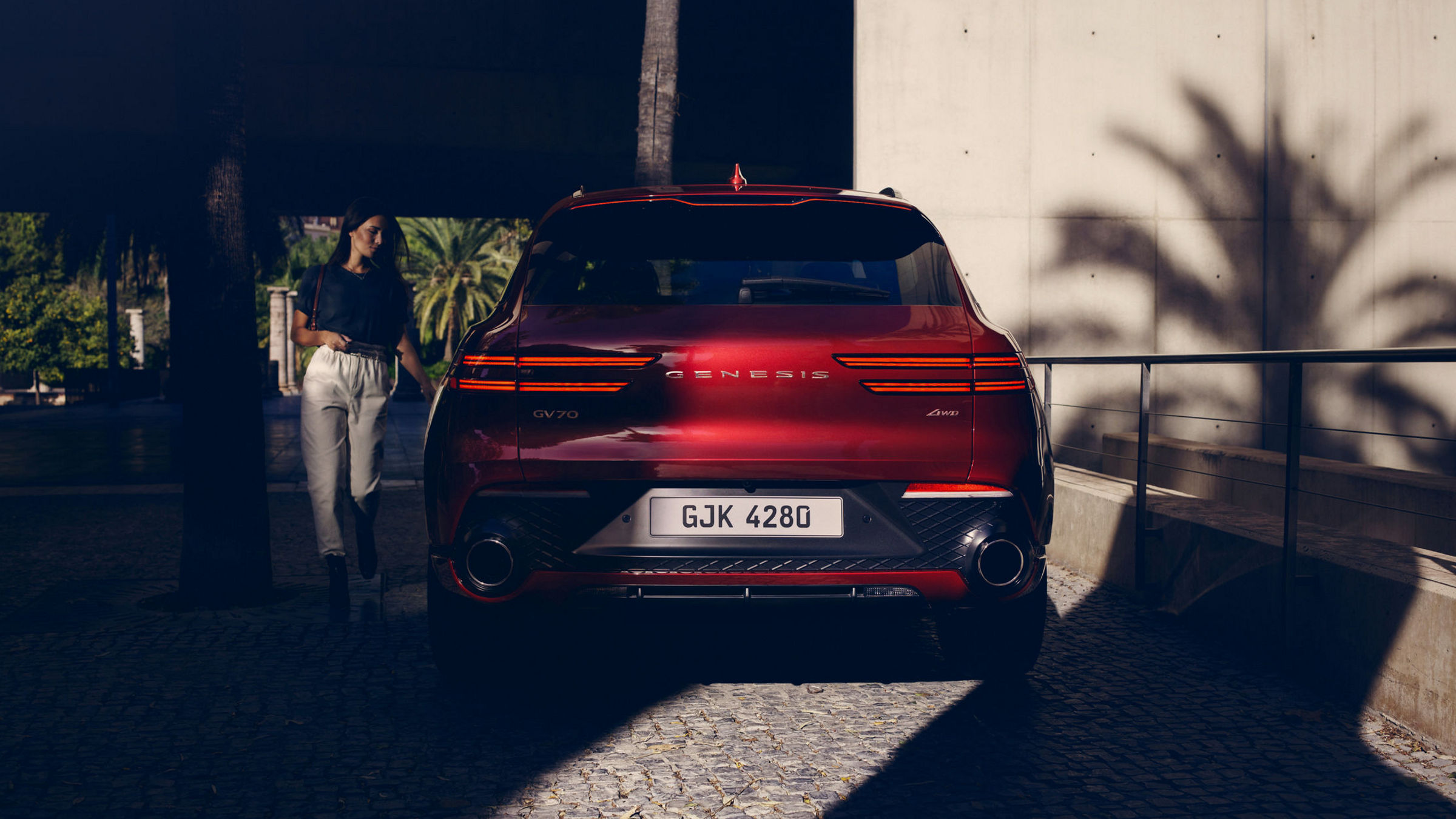 The Genesis GV70 is parked in an urban setting with a modern concrete wall and palm tree shadows, highlighting its sleek rear design and distinctive lighting.