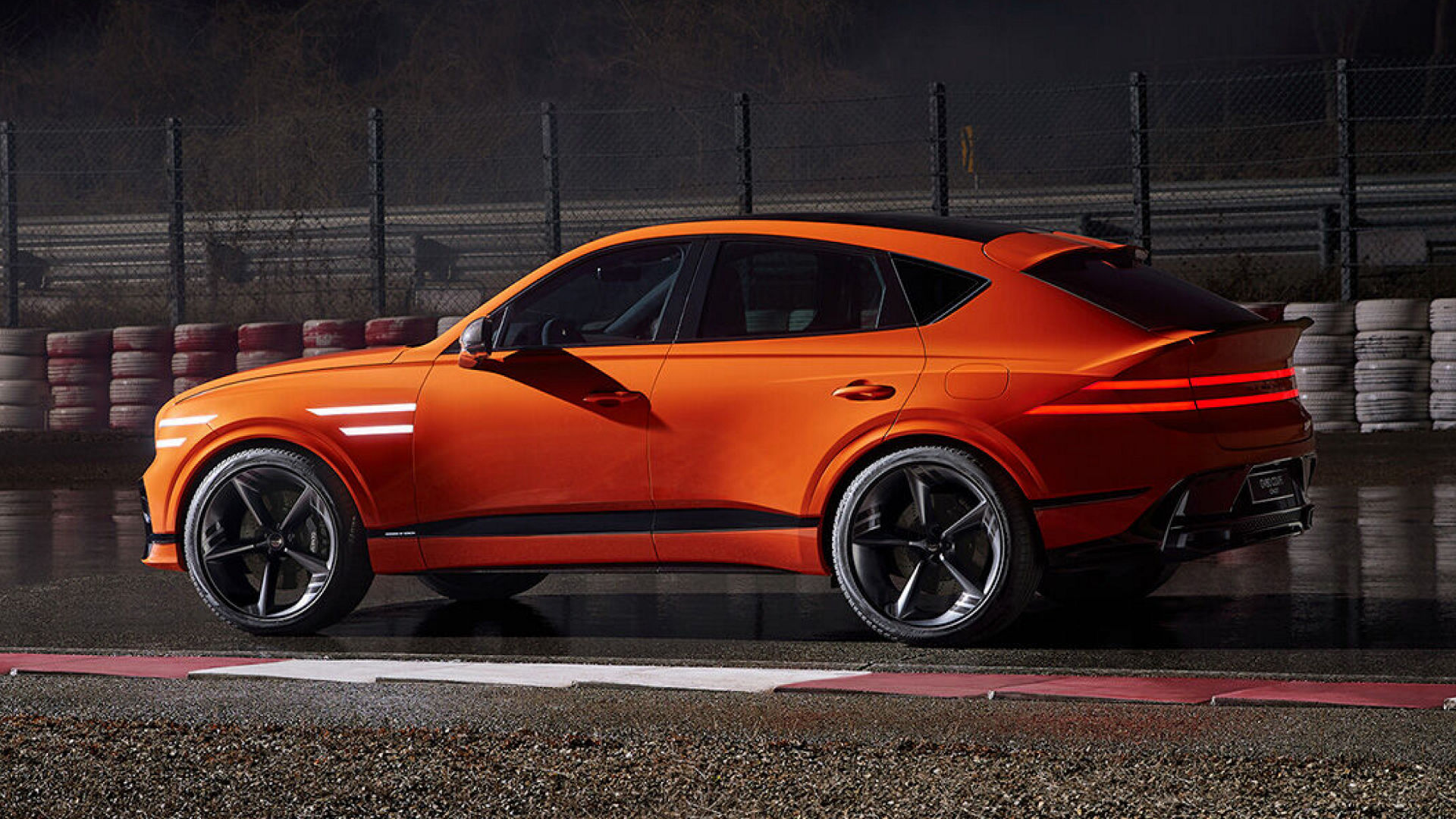 Side view of the Genesis GV80 Coupe in orange, parked on a wet track at night