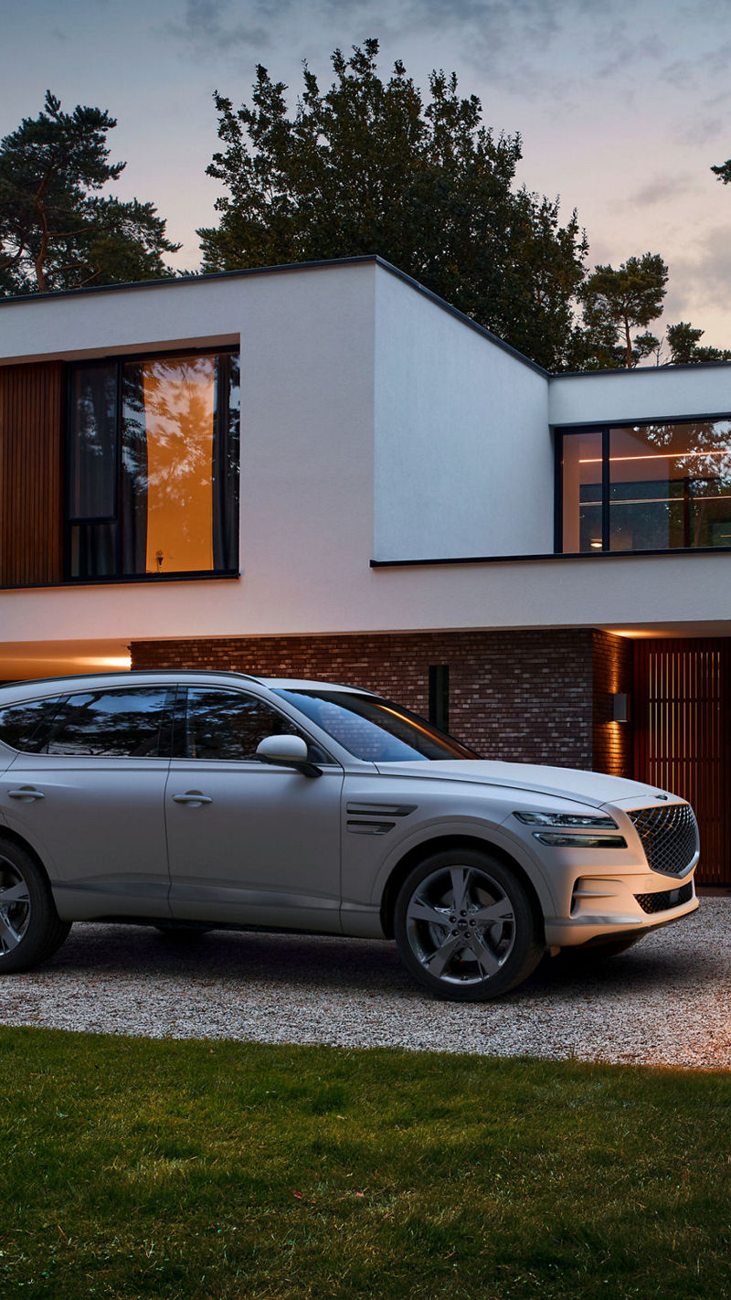 White Genesis GV80 next to a meadow in front of a modern building with several people inside