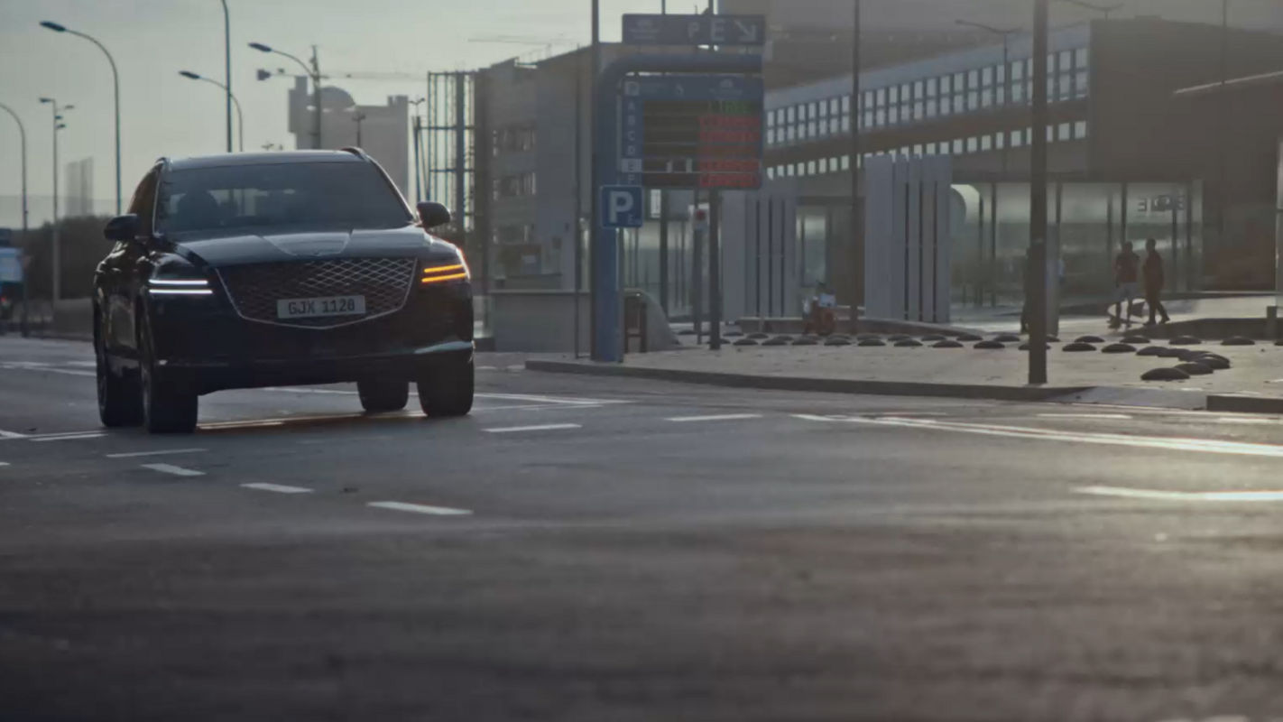 The Genesis Electrified GV80 driving through a modern city street during the evening, with headlights illuminated.