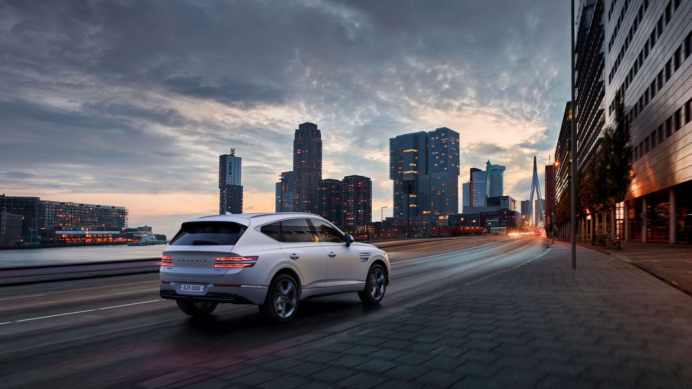 A white Genesis GV80 drives on a city street with skyscrapers in the background.