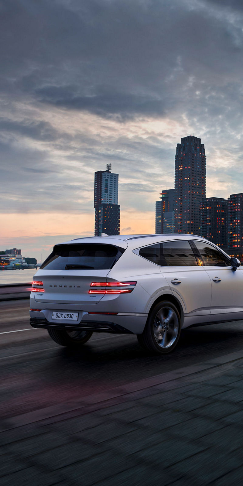 A white Genesis GV80 drives on a city street with skyscrapers in the background.