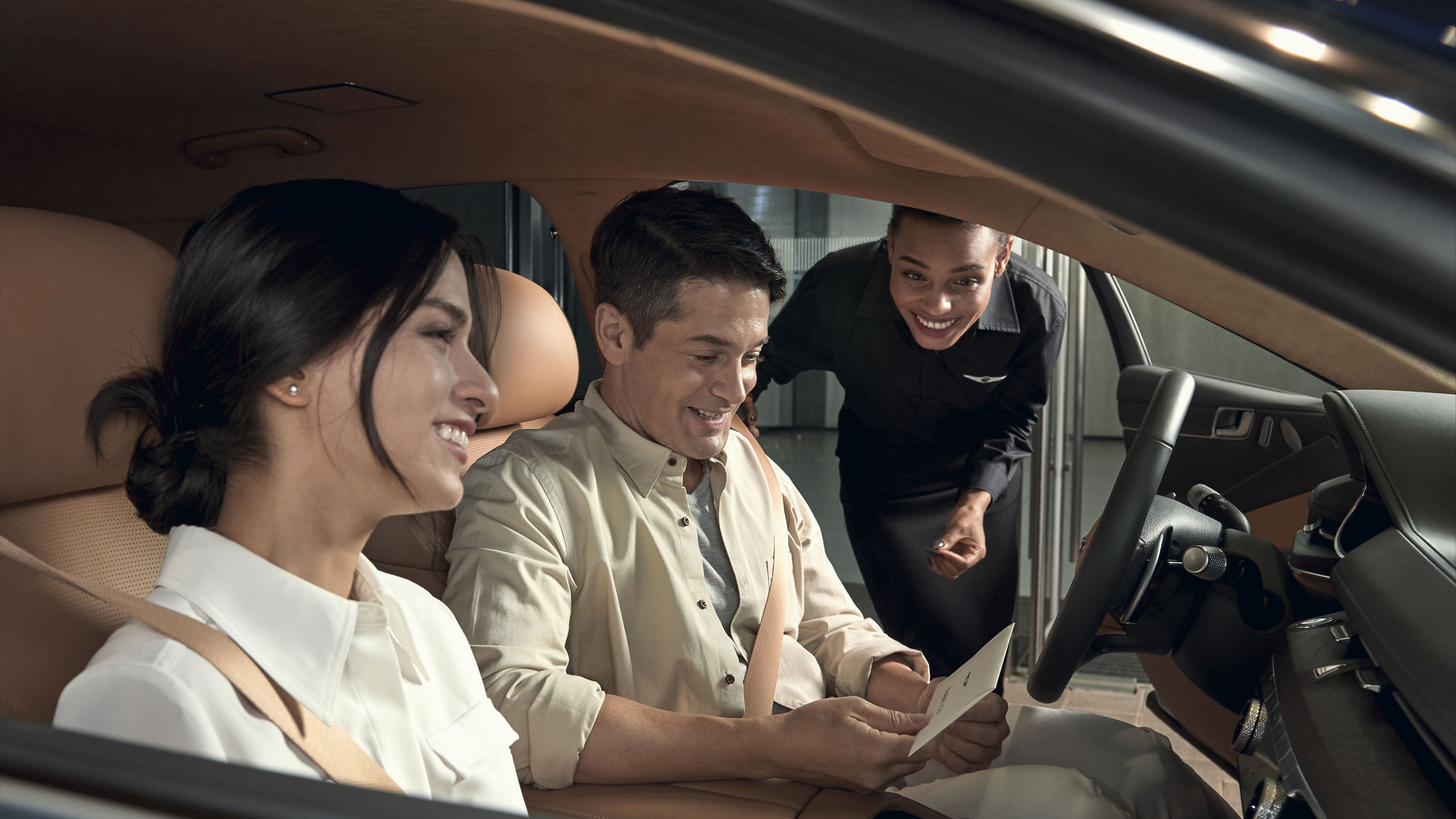 A couple smiling inside a Genesis car as a service advisor hands them documents through the open door.