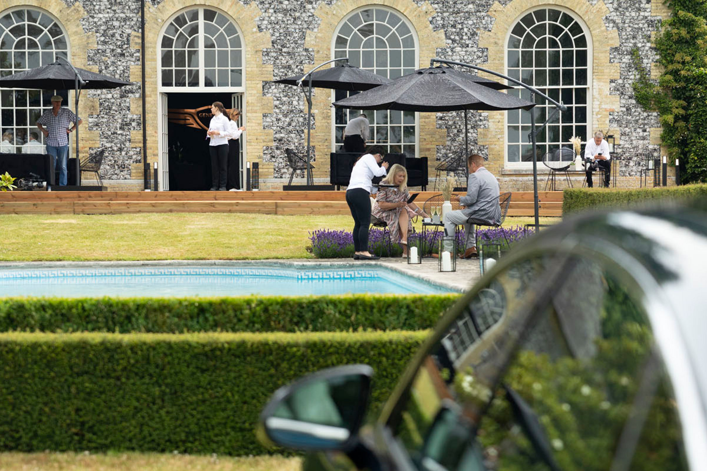 People under parasols in front of a house with a car in the foreground